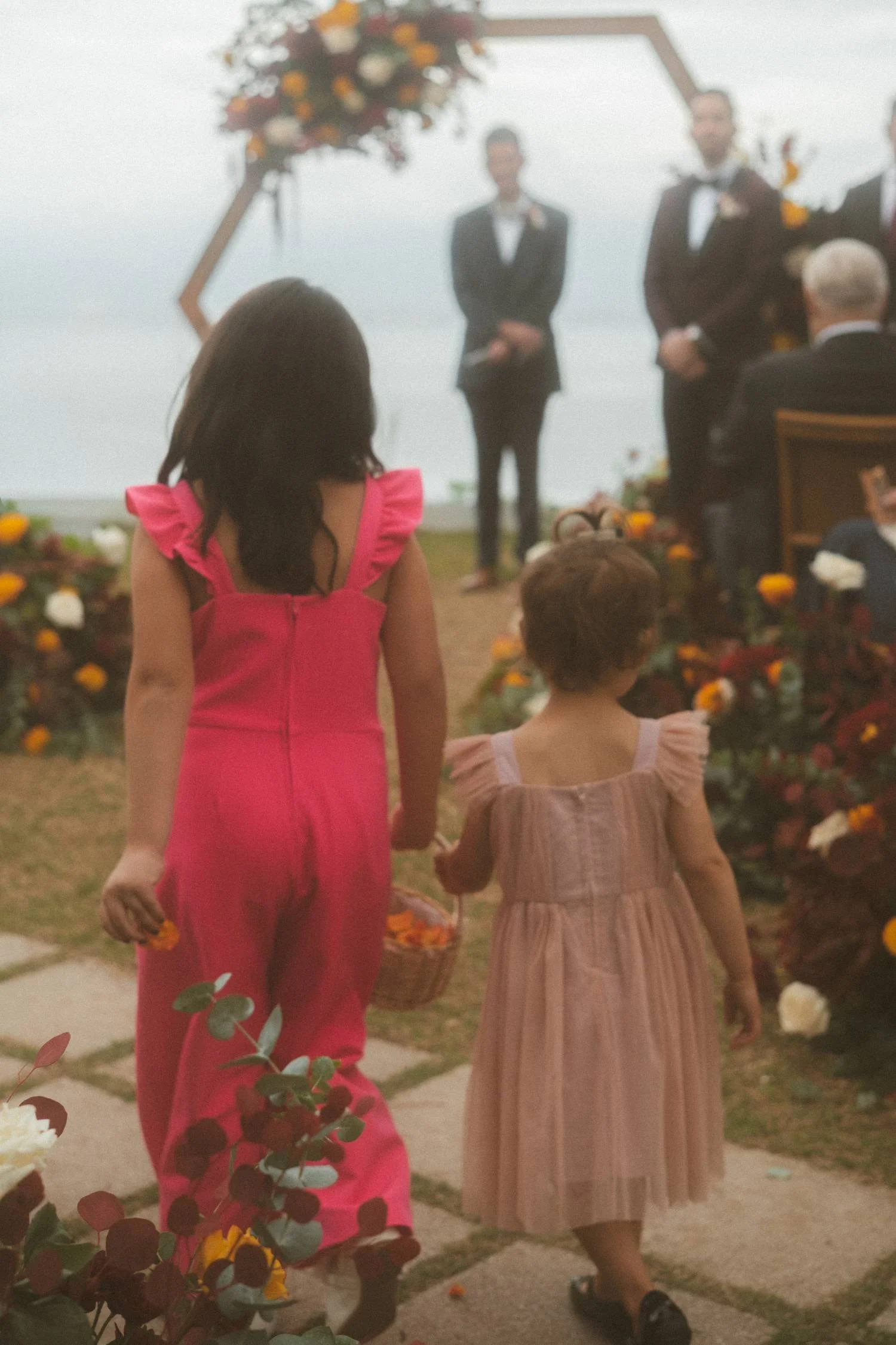 Two young girls walking down an outdoor wedding aisle, one holding a small basket