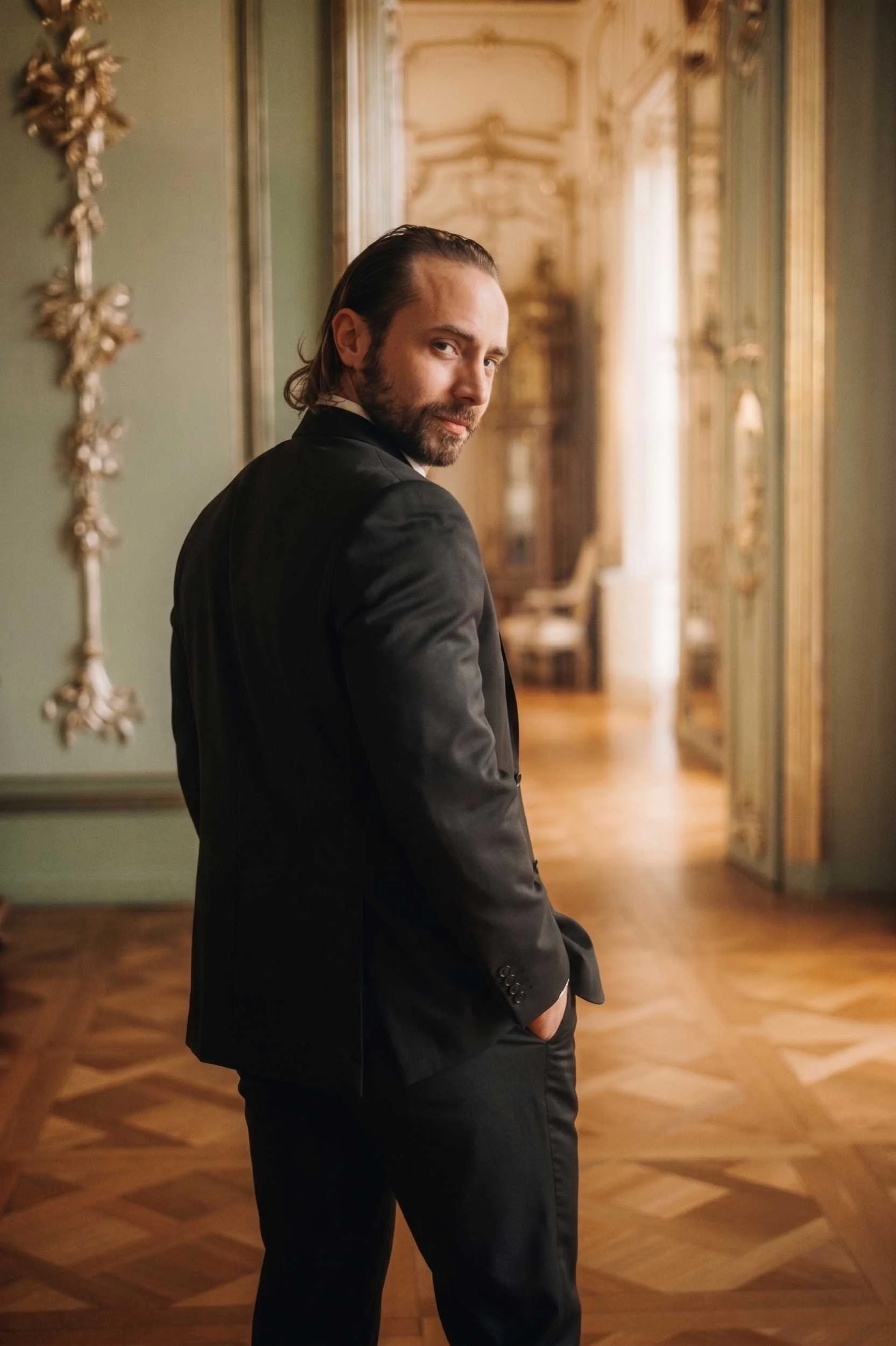 A man in a tailored black suit looking over his shoulder in a luxurious, ornate room.