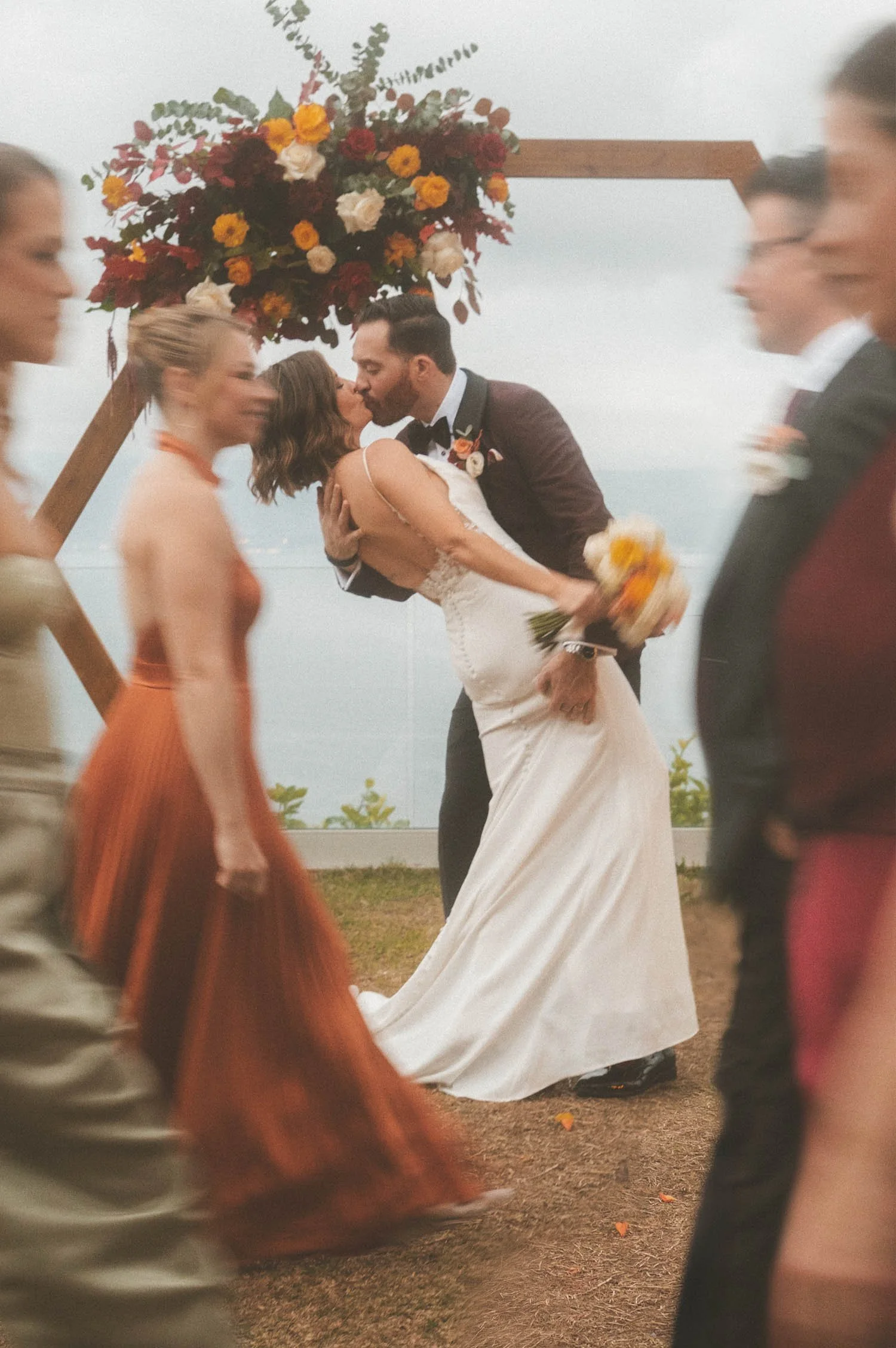 Bride and groom sharing their first kiss during an outdoor wedding ceremony with guests walking past in motion blur