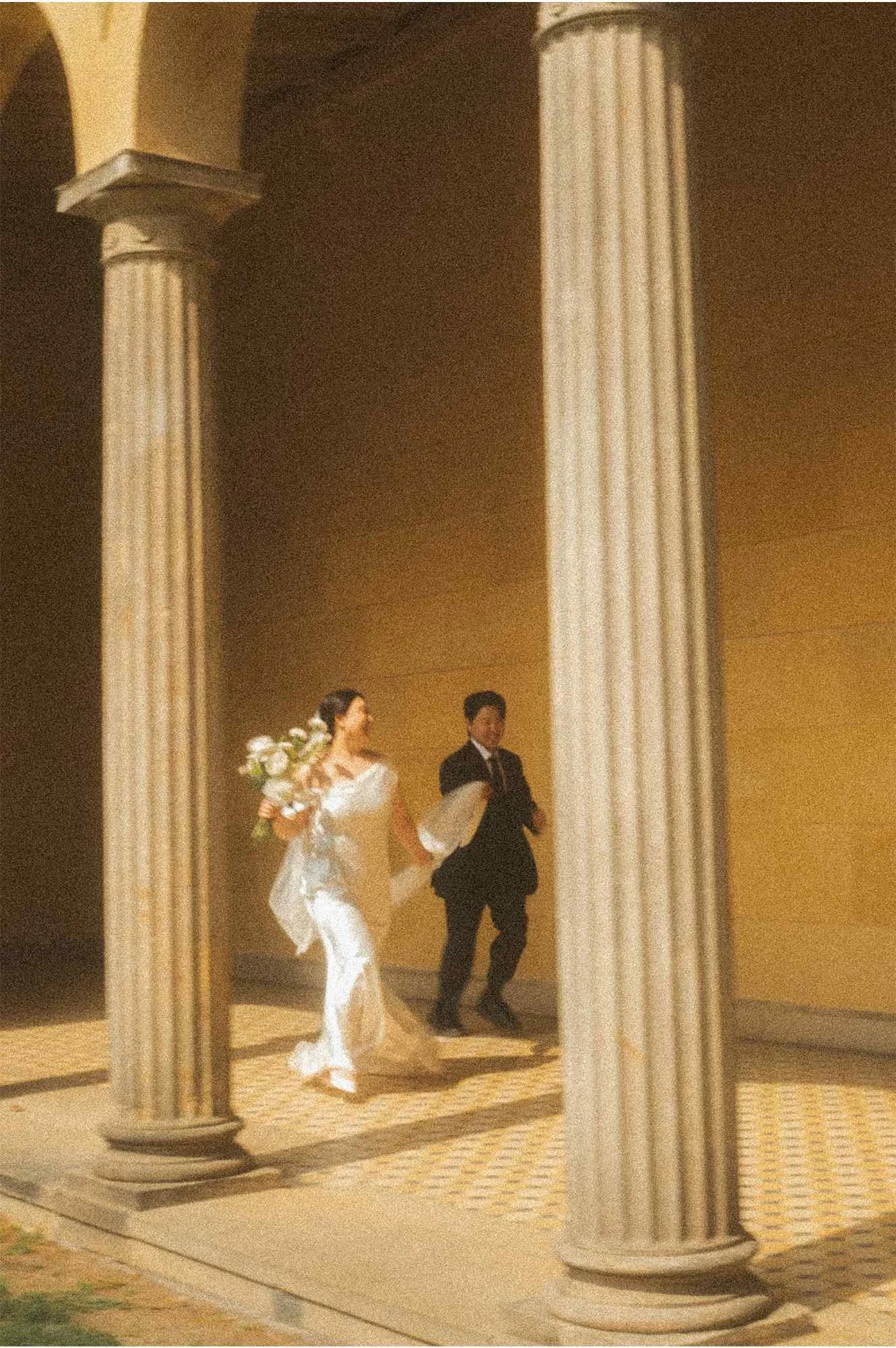 A wide shot of a bride and groom holding hands and running together between large stone columns at a palace in Potsdam during golden hour.