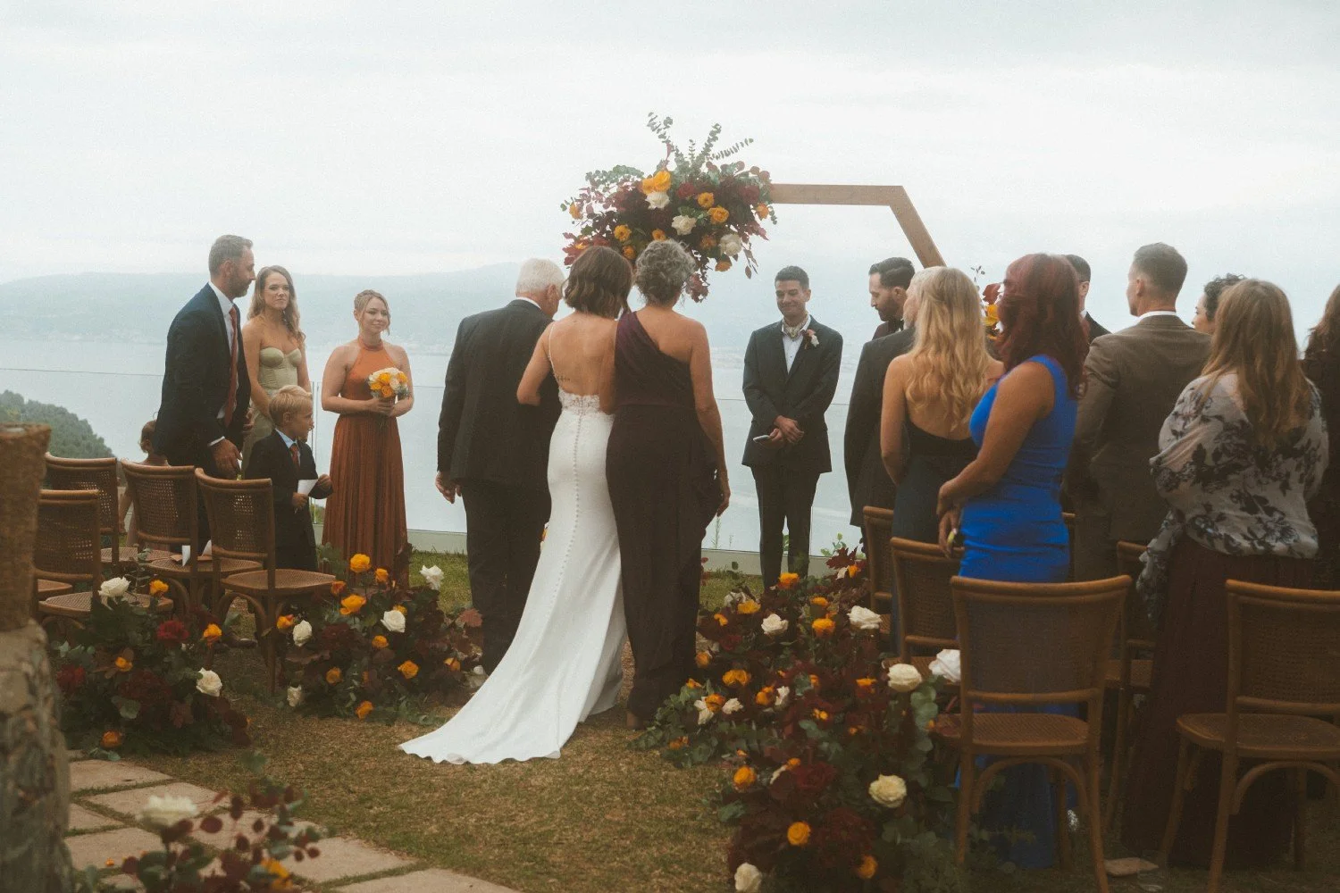 Outdoor lakeside wedding ceremony with bride walking down the aisle toward a floral arch, surrounded by guests