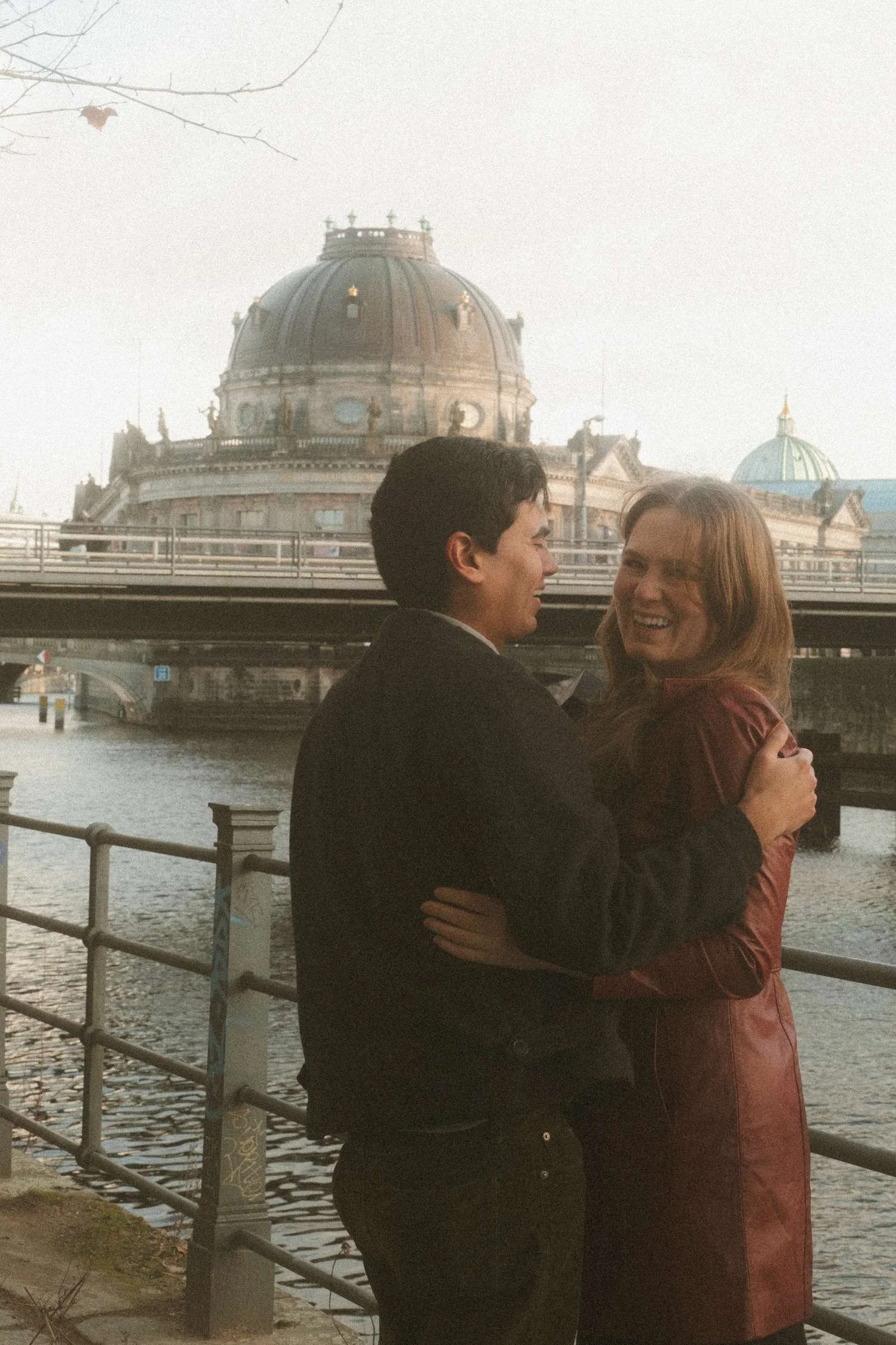 Laughing couple embracing by the river with the Bode Museum dome visible in the background during their Berlin engagement photo session