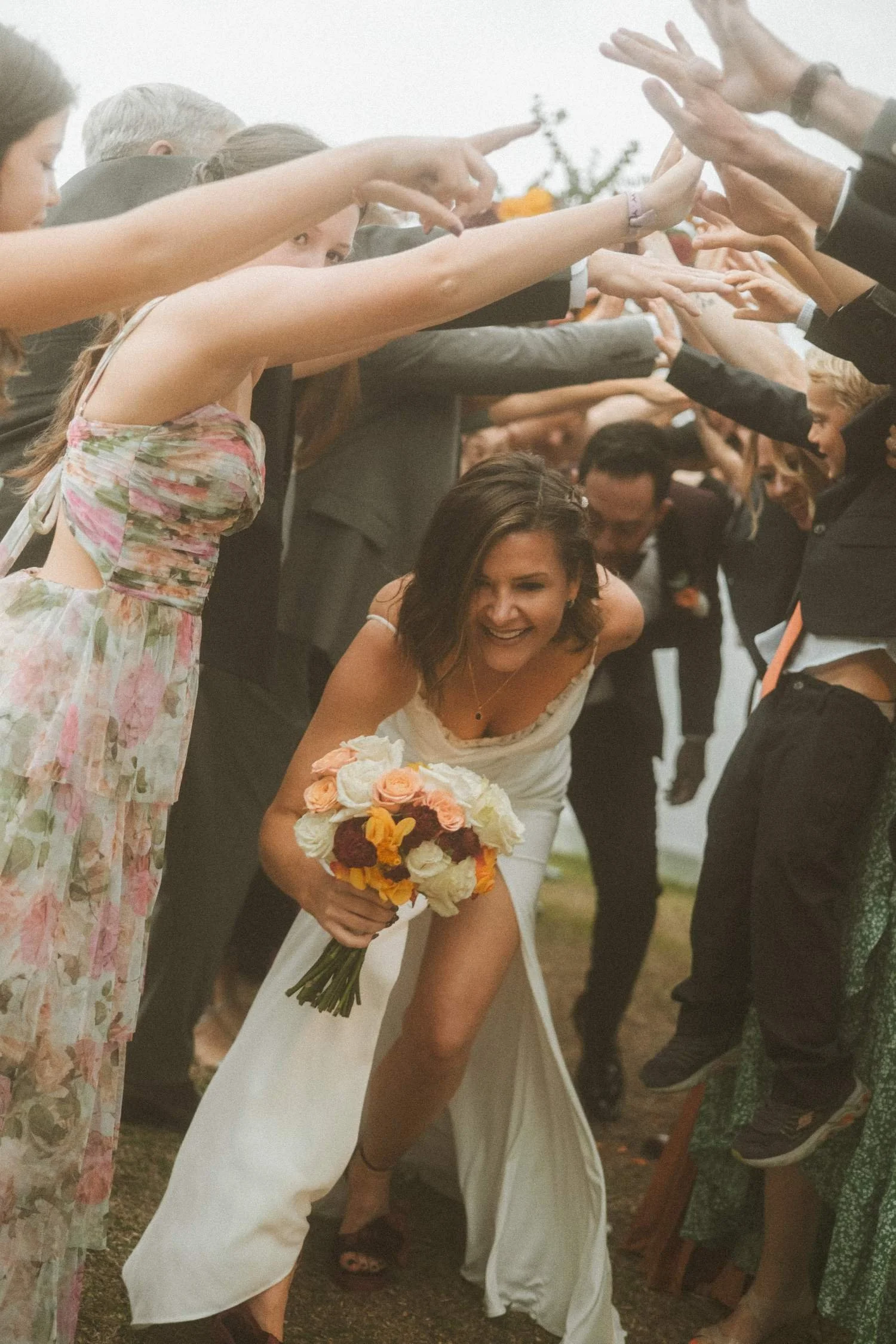 Bride smiling while walking through a tunnel of wedding guests during an outdoor wedding ceremony in Berlin, holding a colorful floral bouquet.