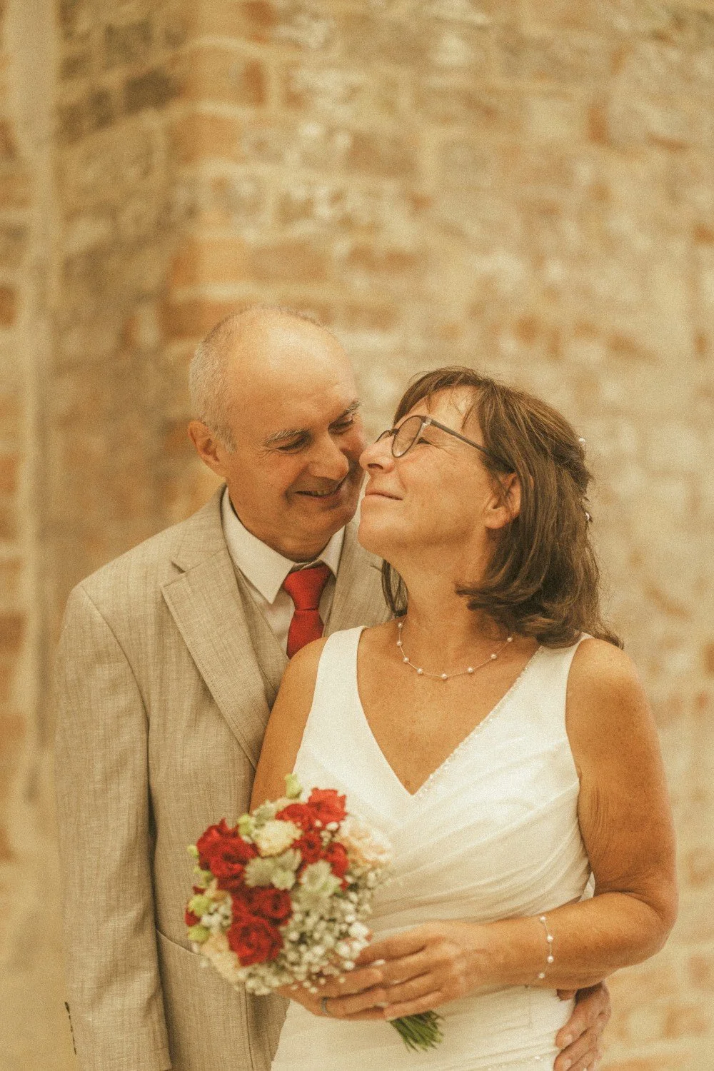 A close-up outdoor portrait of the wedding couple smiling lovingly at each other, with the bride holding a bouquet of red and white flowers.