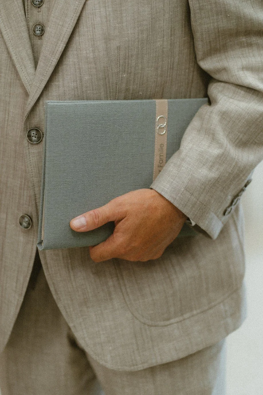 A close-up of a groom wearing a beige suit and holding a green wedding certificate folder.