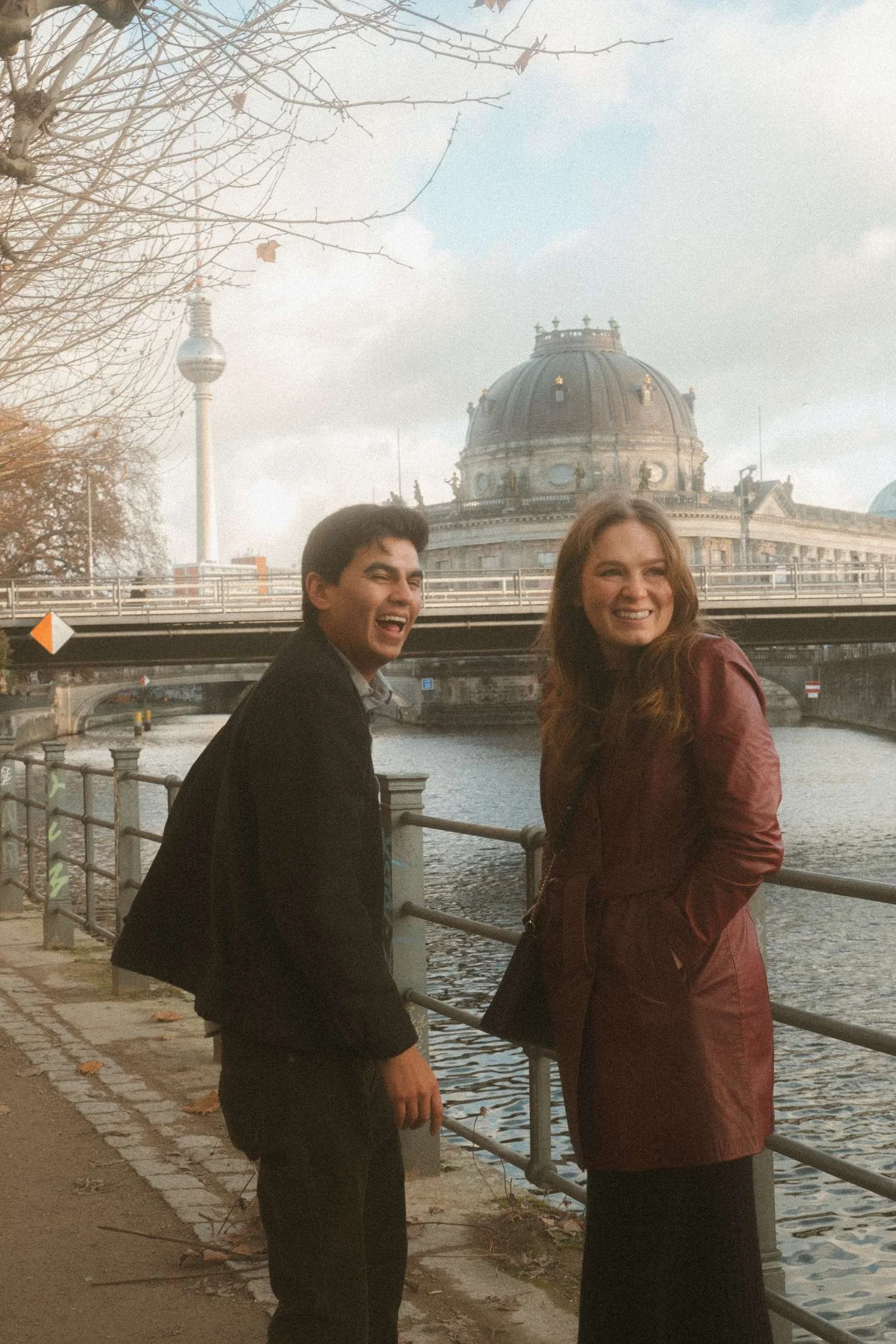 Laughing couple standing by the Spree River with the Berlin TV Tower and Bode Museum dome in the background during their engagement photo session