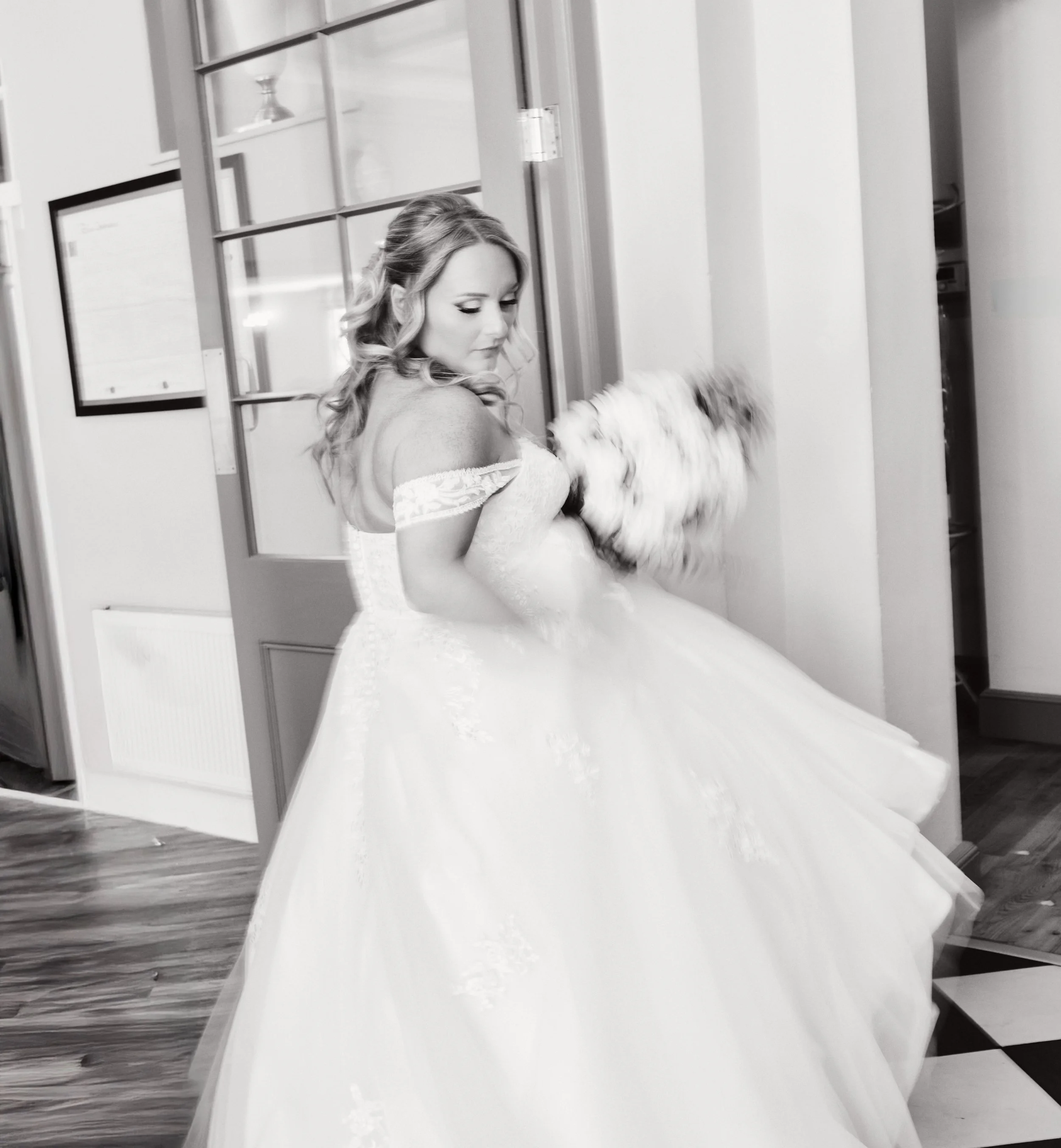 Black and white photo of a bride in an off-shoulder wedding dress, spinning or twirling in an indoor space with wooden and tiled floors, holding a bouquet of flowers.