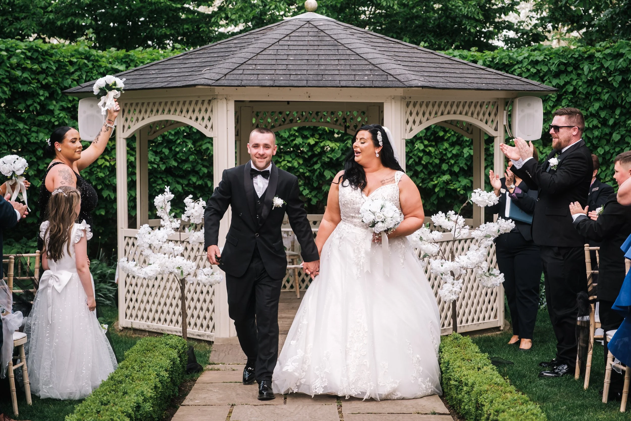 Newlywed couple walking hand in hand down the aisle during their wedding ceremony, surrounded by friends and family clapping and celebrating, outdoors with greenery and a gazebo in the background.