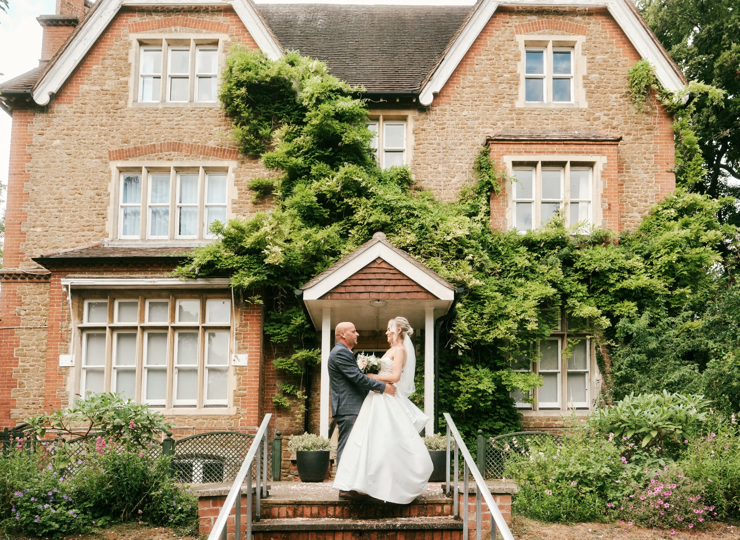A bride and groom standing on front steps of a brick house decorated with green vines, holding hands and looking at each other.