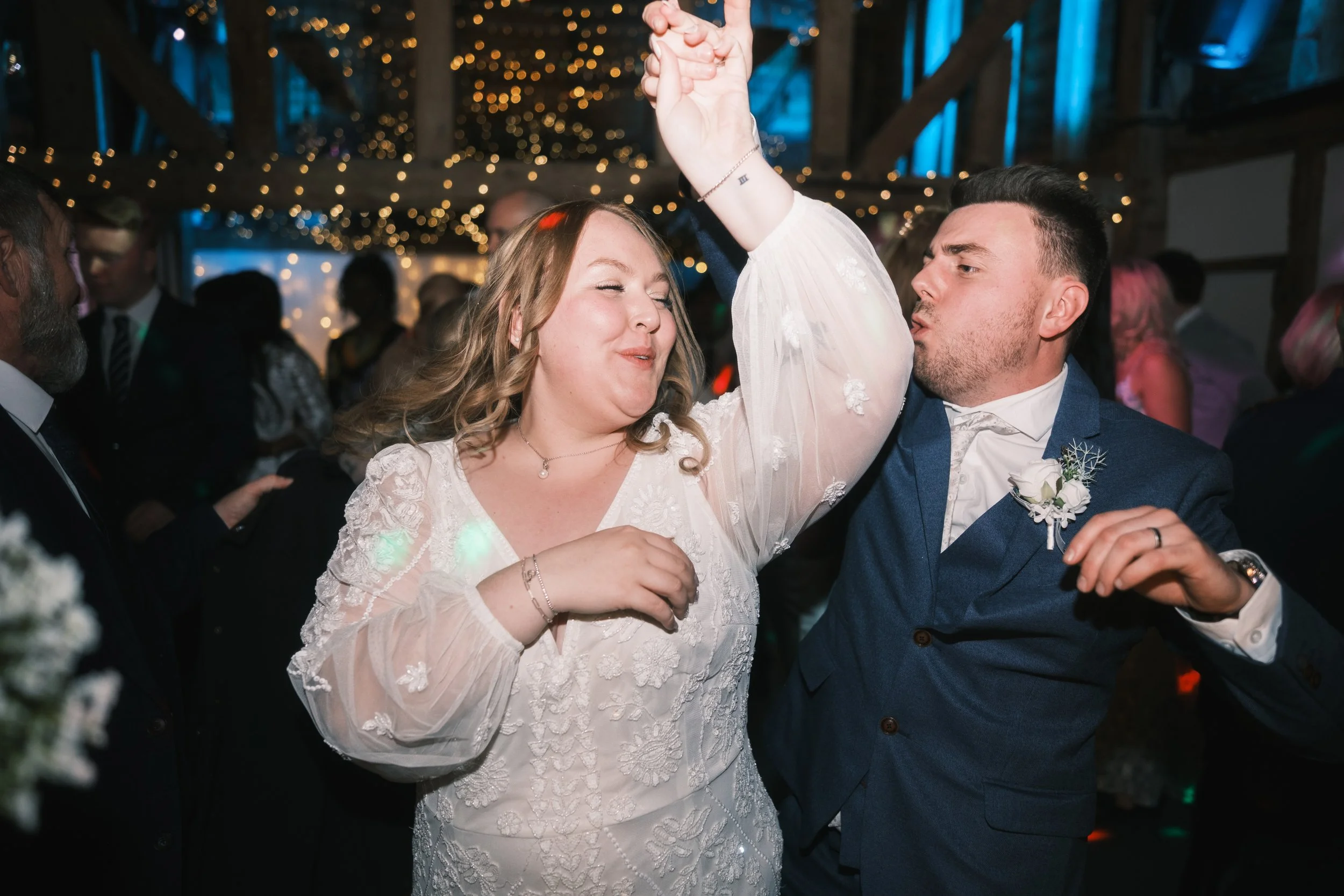 A bride and groom dancing at a wedding reception, with bride wearing a lace dress and groom in a navy suit, in a decorated venue with string lights.