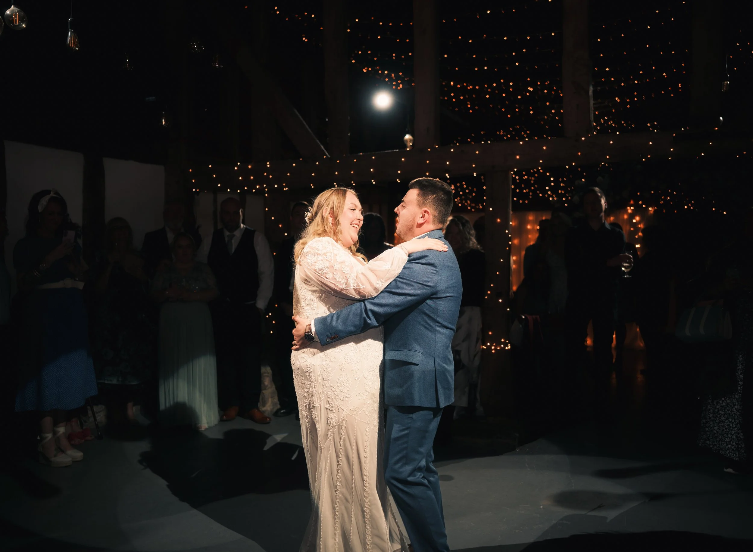 A bride and groom sharing their first dance at a wedding reception, surrounded by guests, under warm string lights in a rustic barn