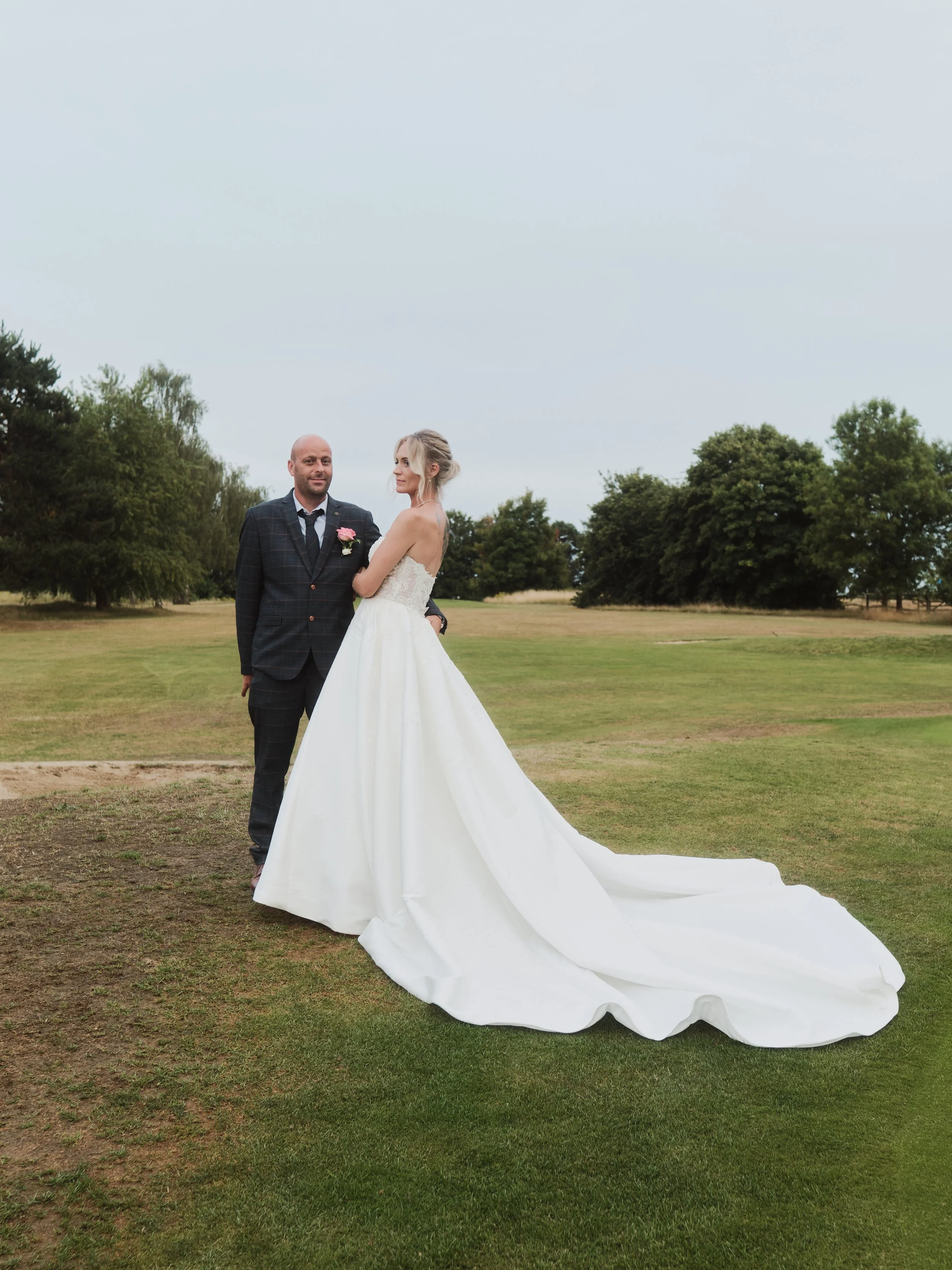 Bride in white wedding gown and groom in dark suit standing on golf course with green trees and cloudy sky in background.