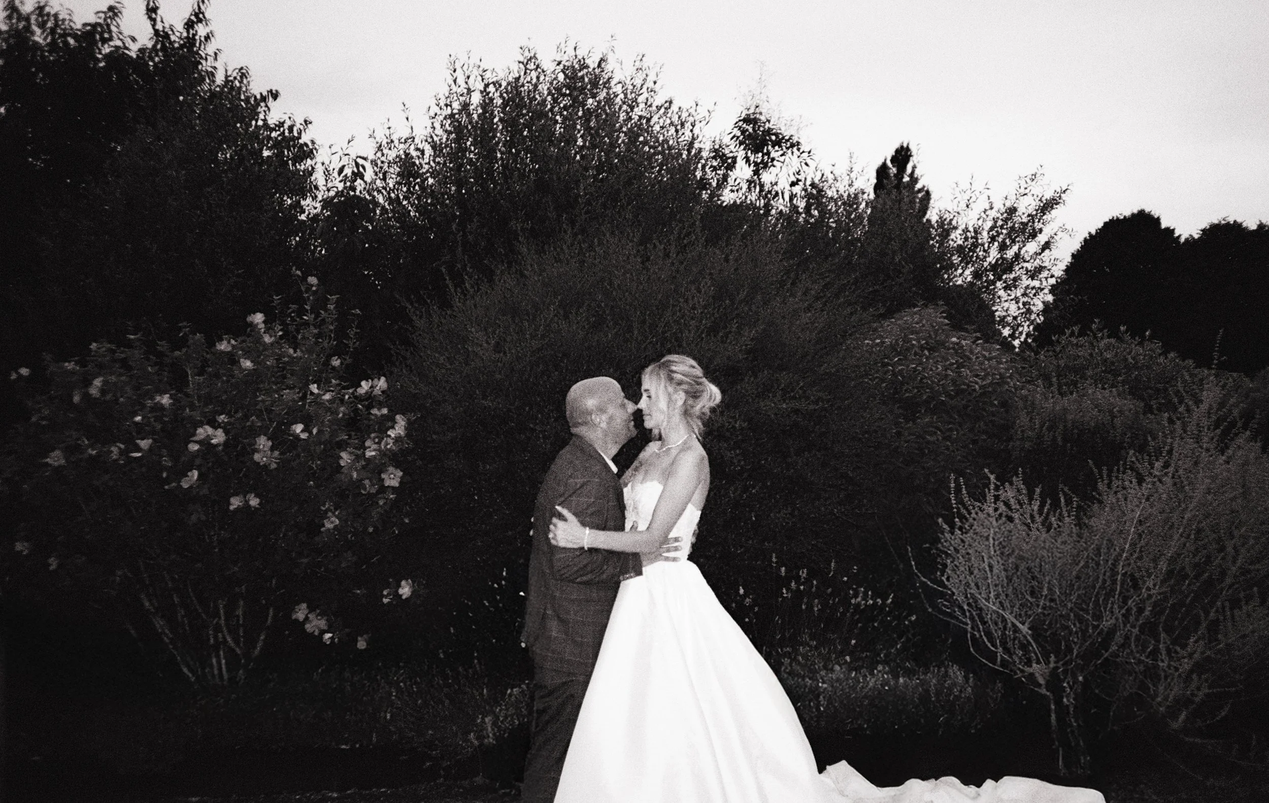 A couple in wedding attire sharing a romantic moment outdoors with trees and bushes in the background.