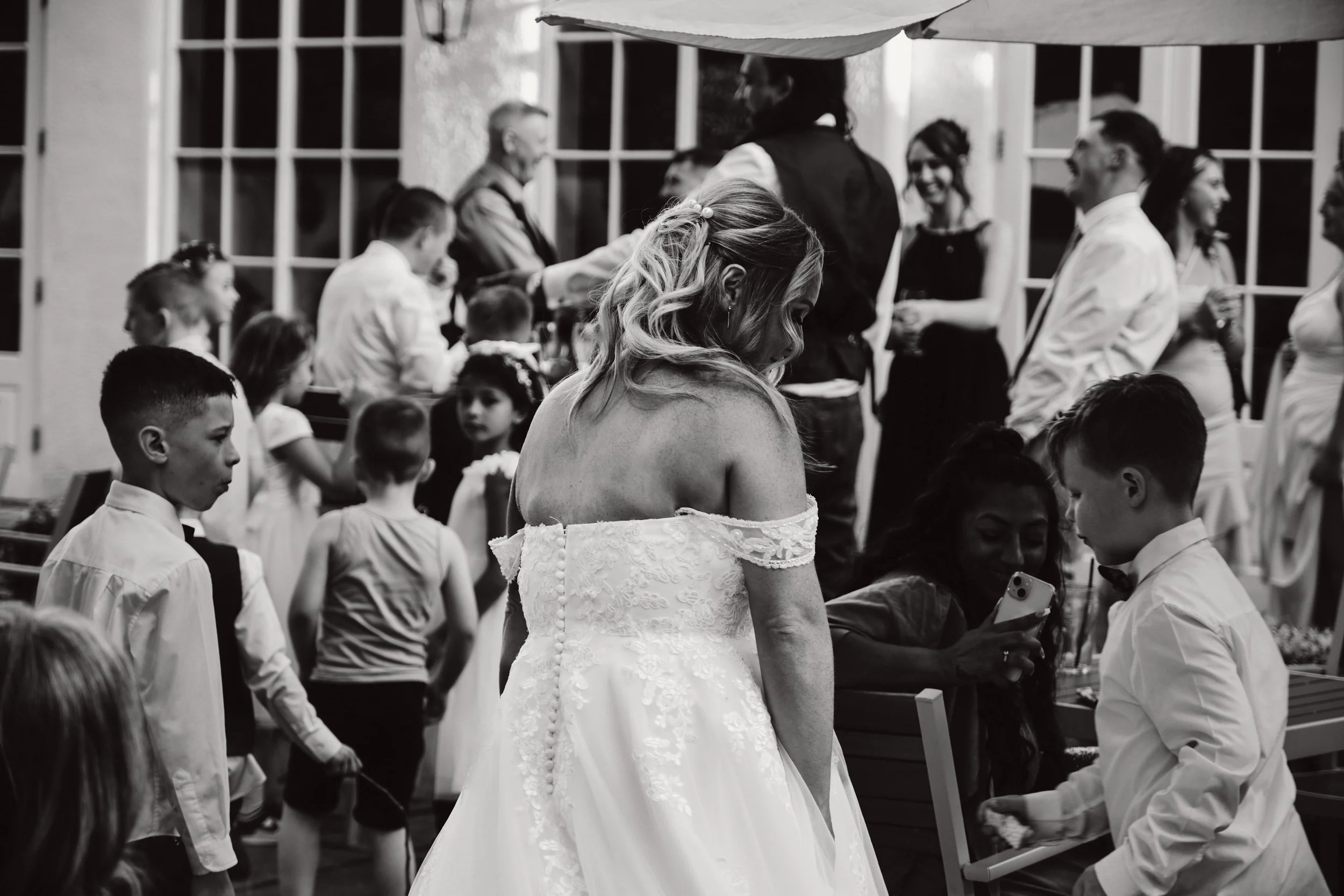 Black and white photo of a wedding reception with adults and children. A woman in a wedding dress is in the foreground, turned away, with children and other guests in the background.