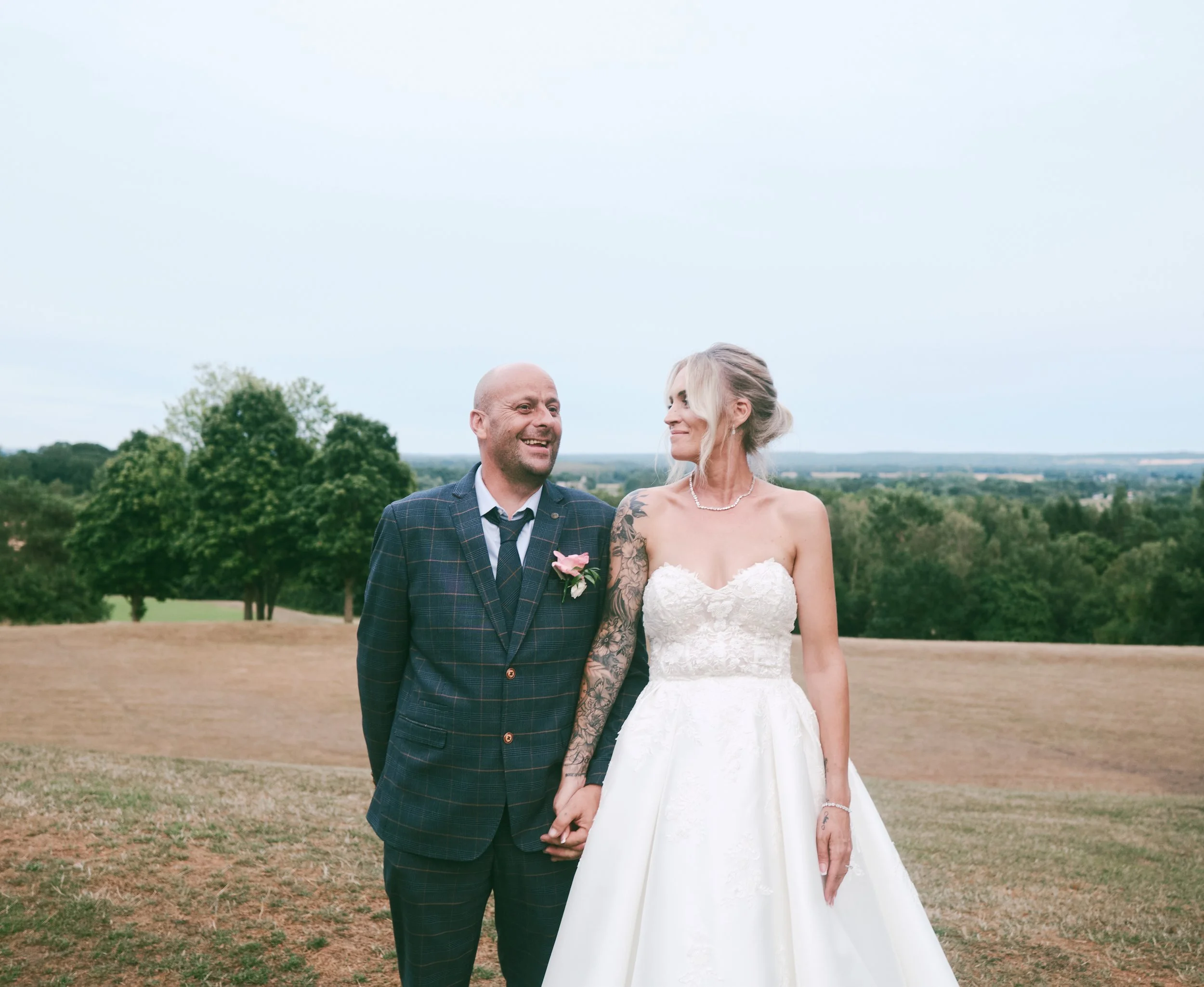 A married couple on their wedding day standing outdoors in a grassy field, holding hands and smiling at each other with a backdrop of trees and a cloudy sky.