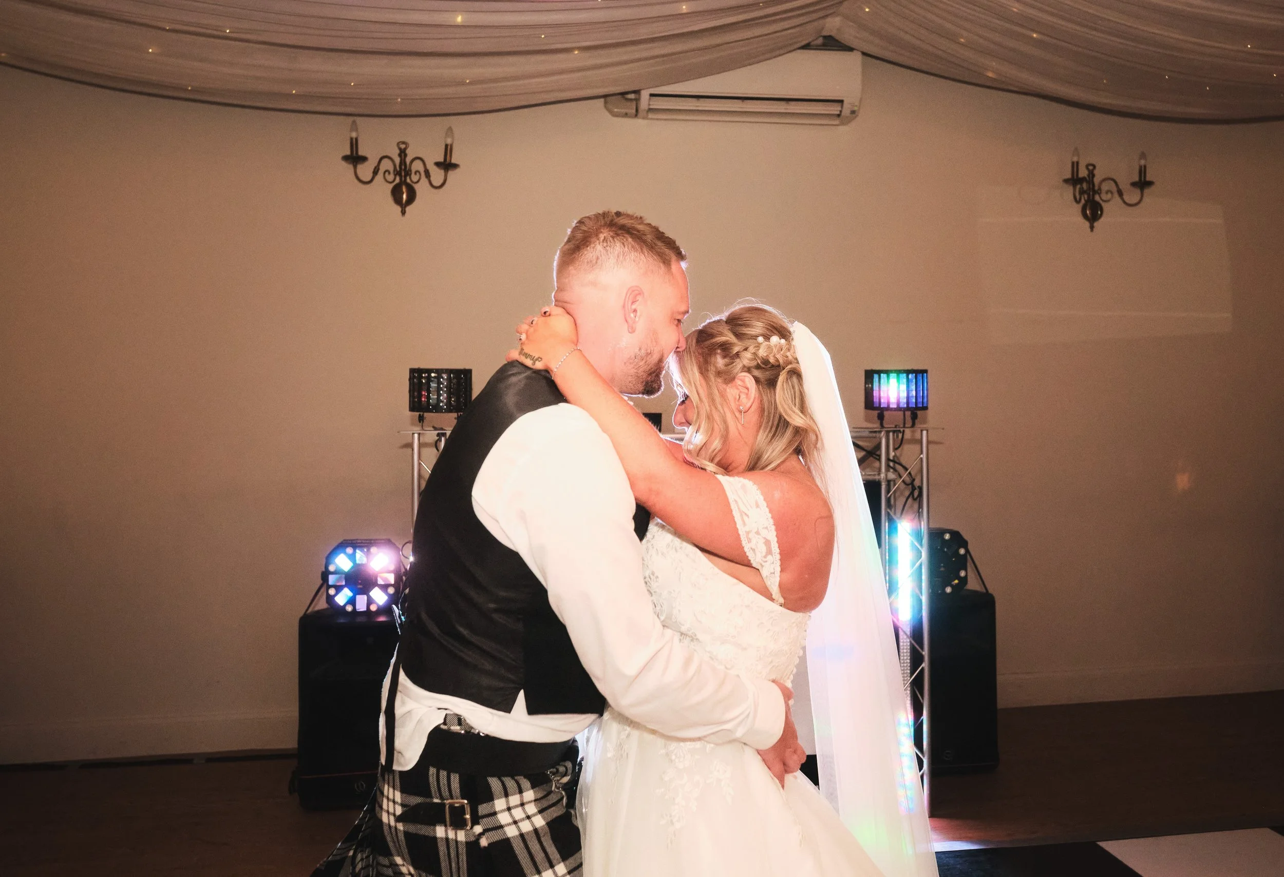 A bride and groom share a dance at their wedding reception, standing closely with foreheads touching in a dimly lit room with uplighting and decorative chandeliers.