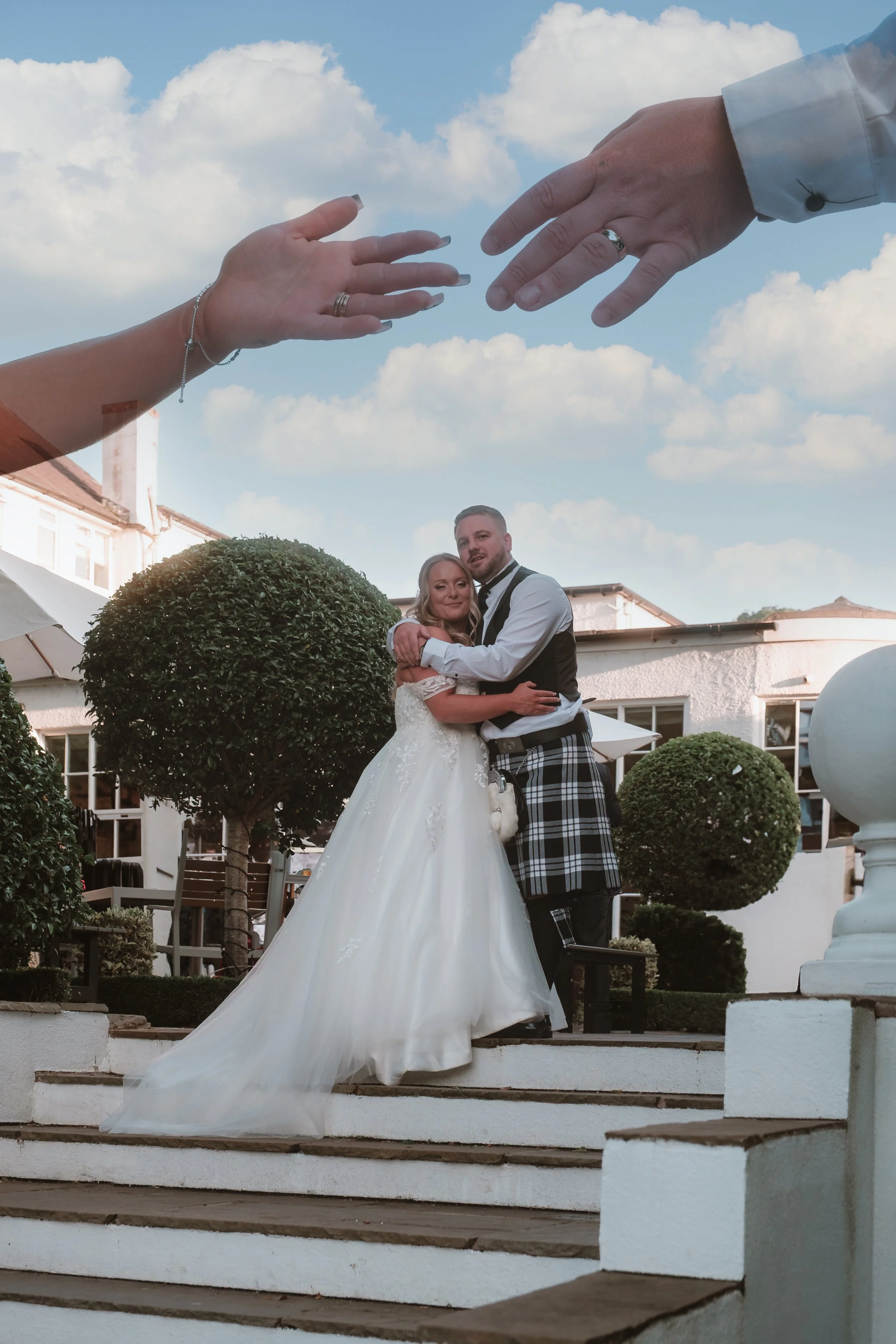 A bride and groom embracing on outdoor wedding stairs, with large staged hands reaching toward each other above them, set against a blue sky with clouds.