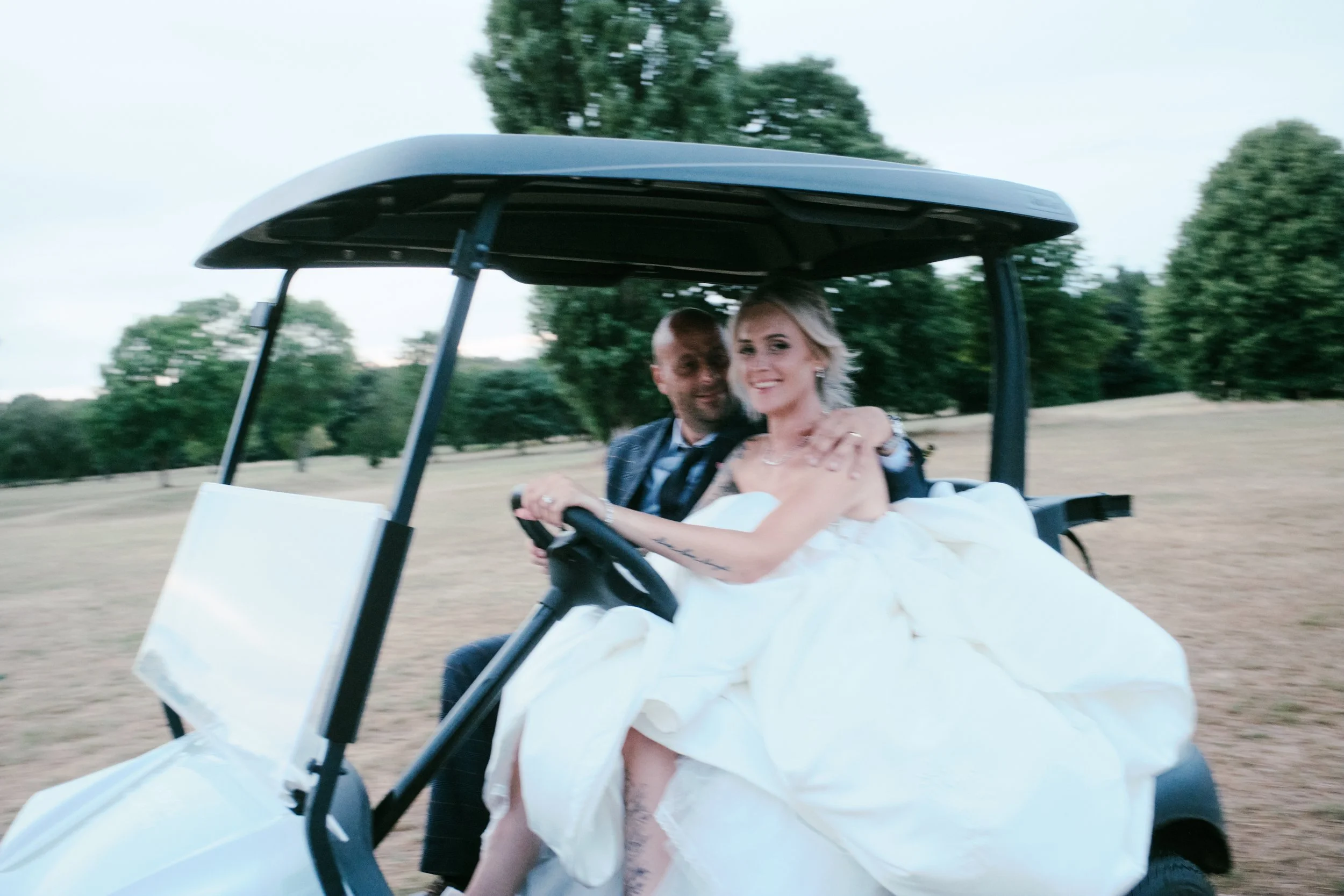 A bride and groom sitting in a golf cart on a golf course, with trees and open land in the background. The bride is wearing a white wedding dress and the groom is in a suit, both smiling.