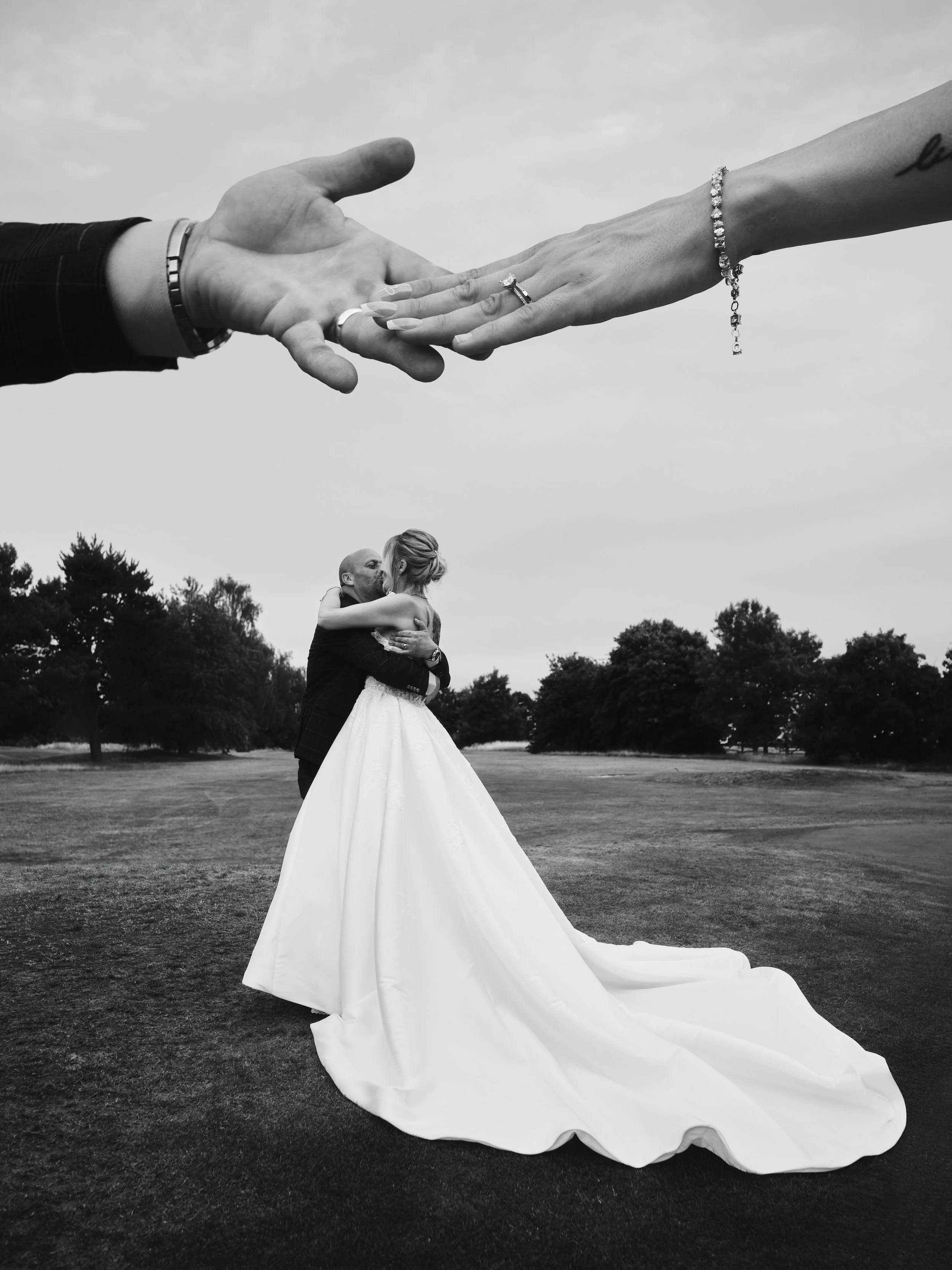 A wedding couple hugging outdoors underneath a large giant pair of hands holding a ring and a bracelet, with trees and sky in the background.