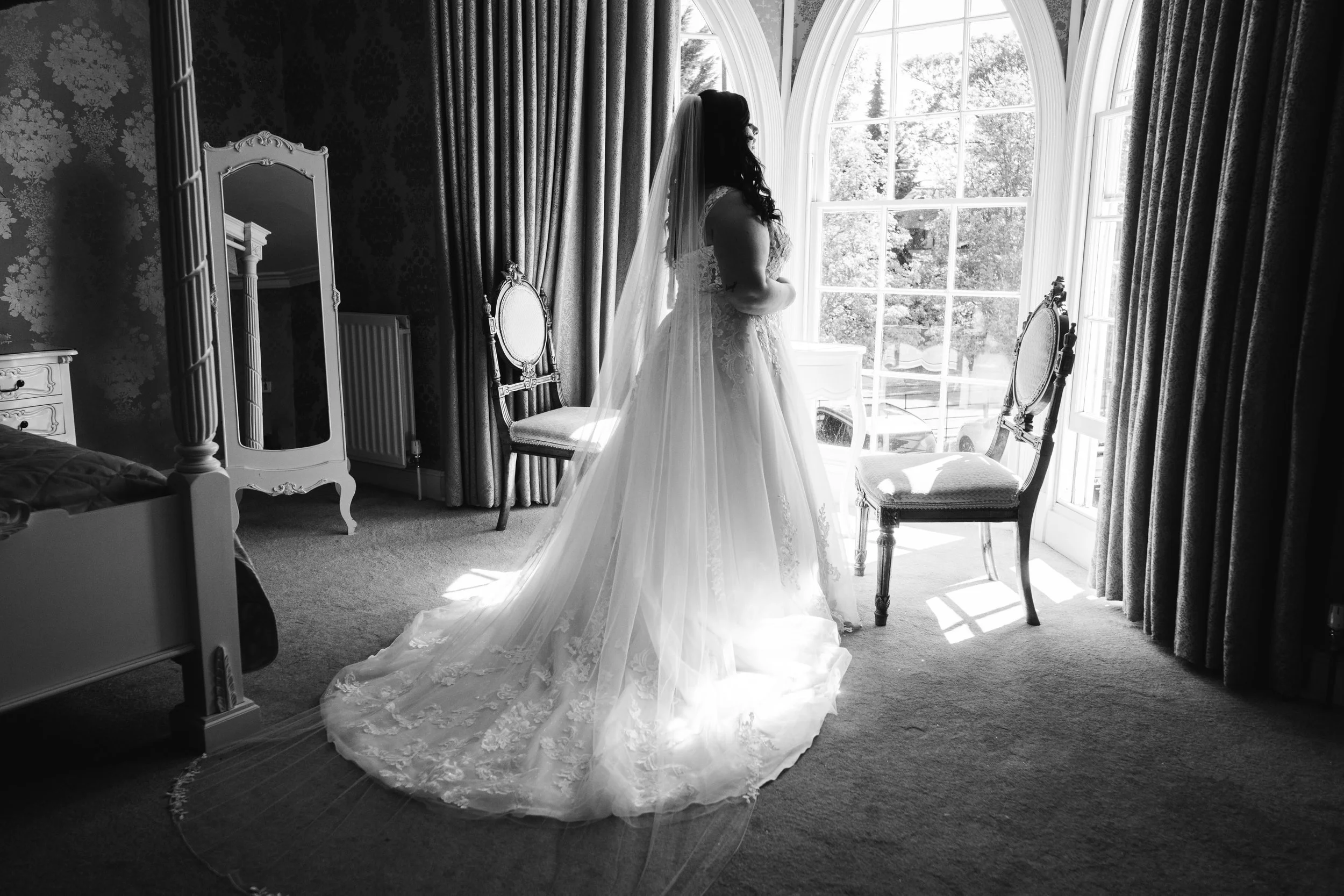 A bride in a wedding dress and veil standing in a bright room with large arched window, ornate chairs, and vintage furniture. The bride is facing the window with sunlight illuminating her silhouette.