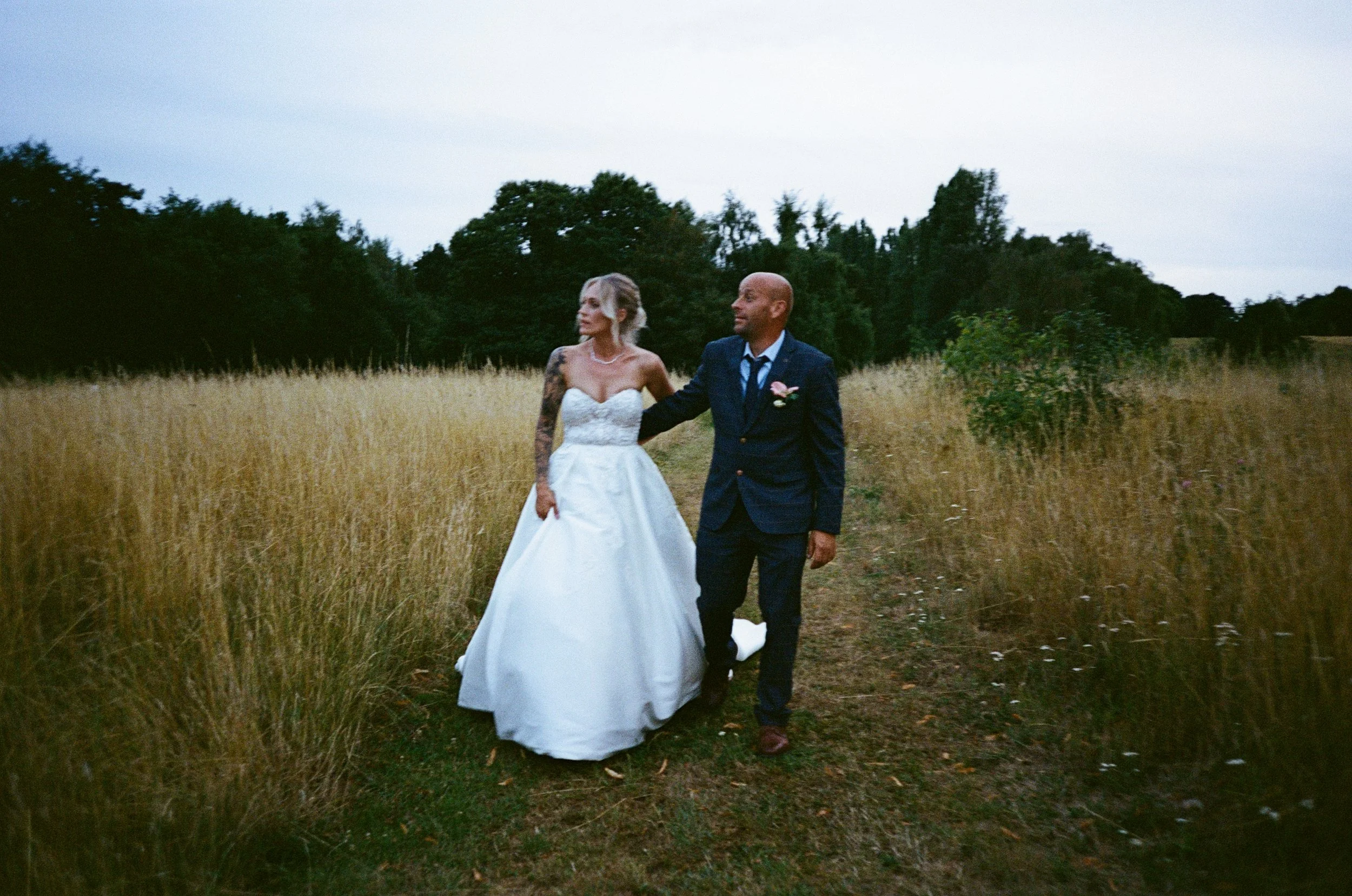 A bride and groom walking together on a grassy dirt path in a field with tall grass, trees in the background, and an overcast sky.