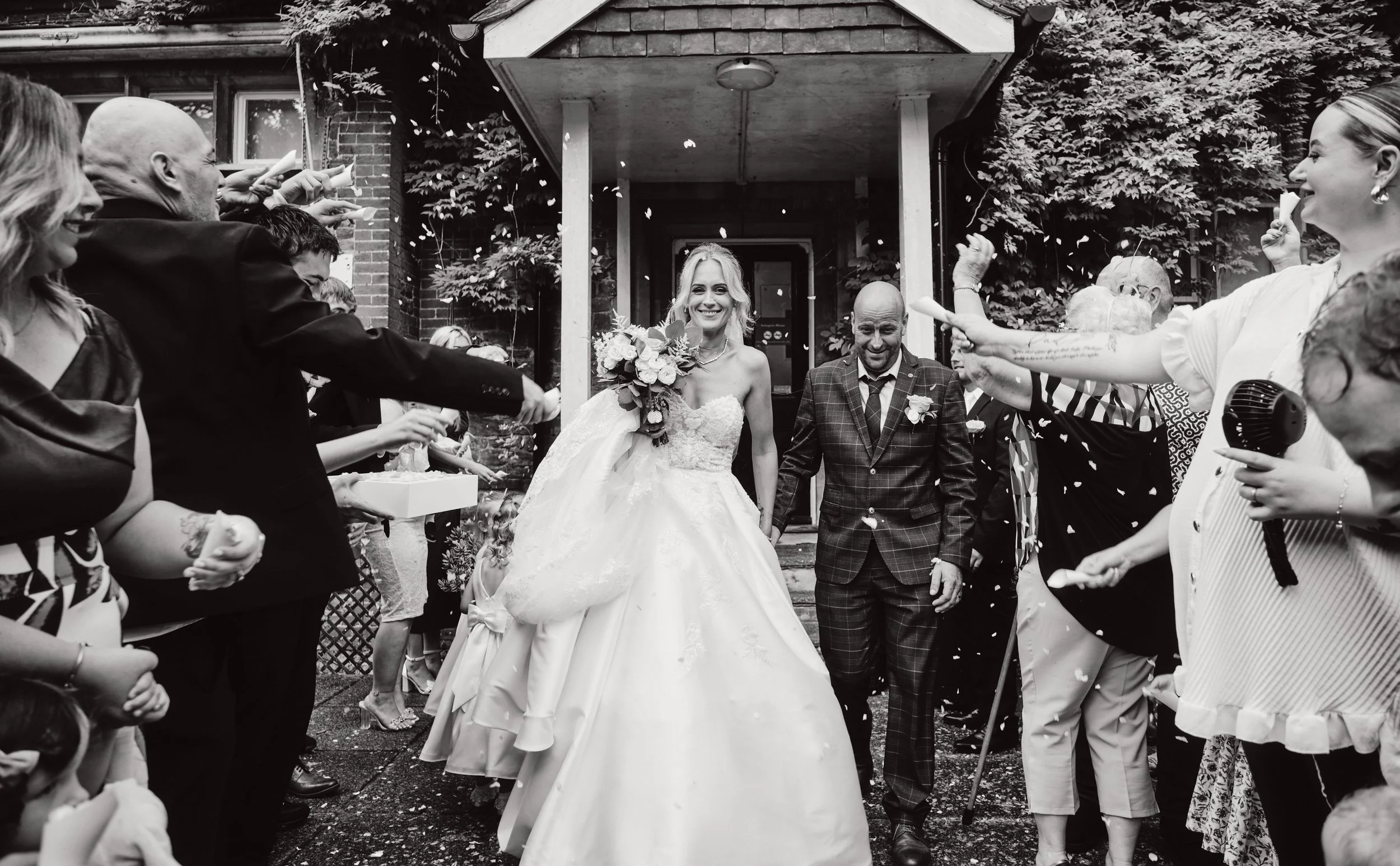 A bride and groom smiling as they walk through a crowd of guests celebrating with confetti outside a building.