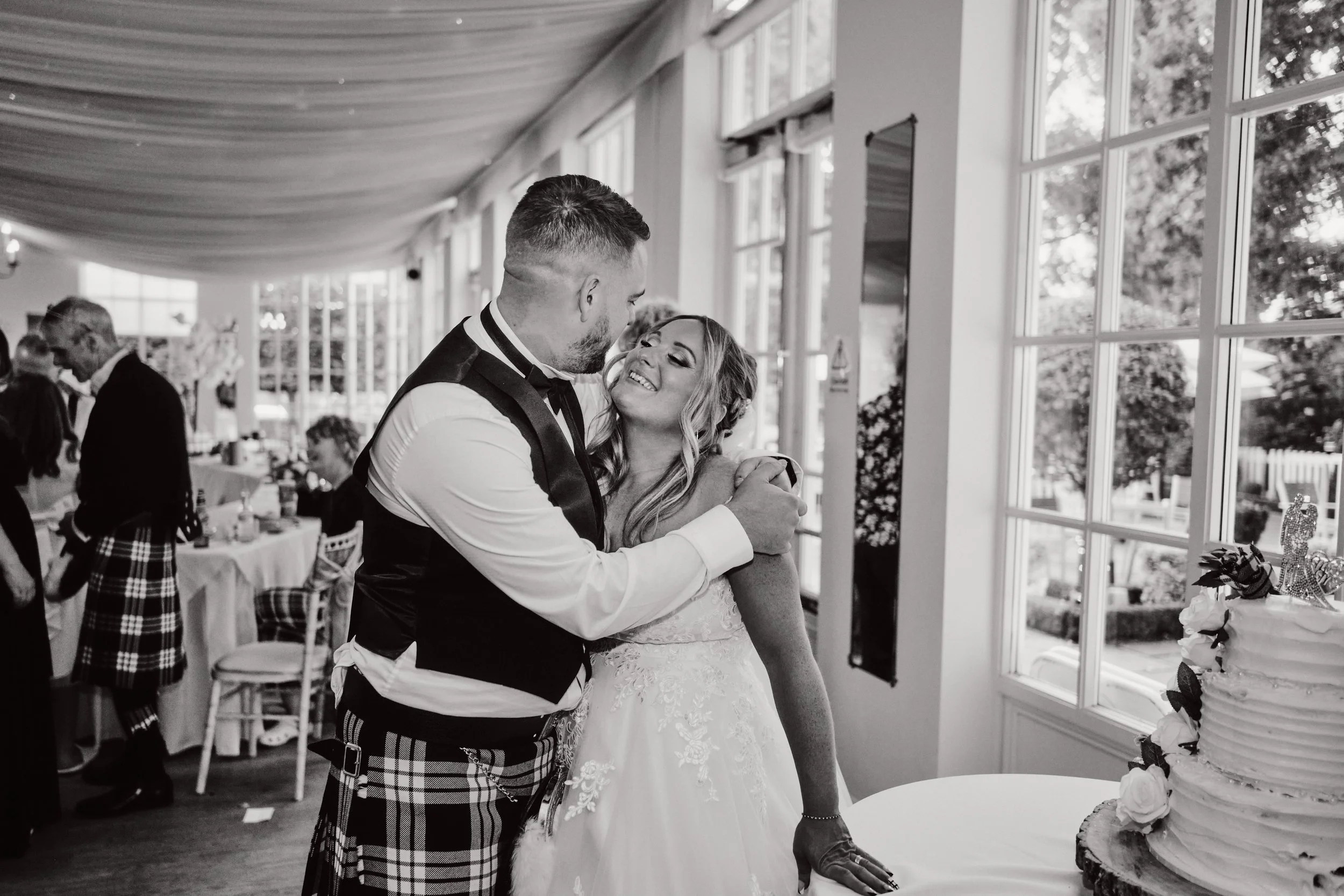 A couple, dressed in wedding attire, sharing a loving moment at their wedding reception inside a bright, decorated venue with large windows.