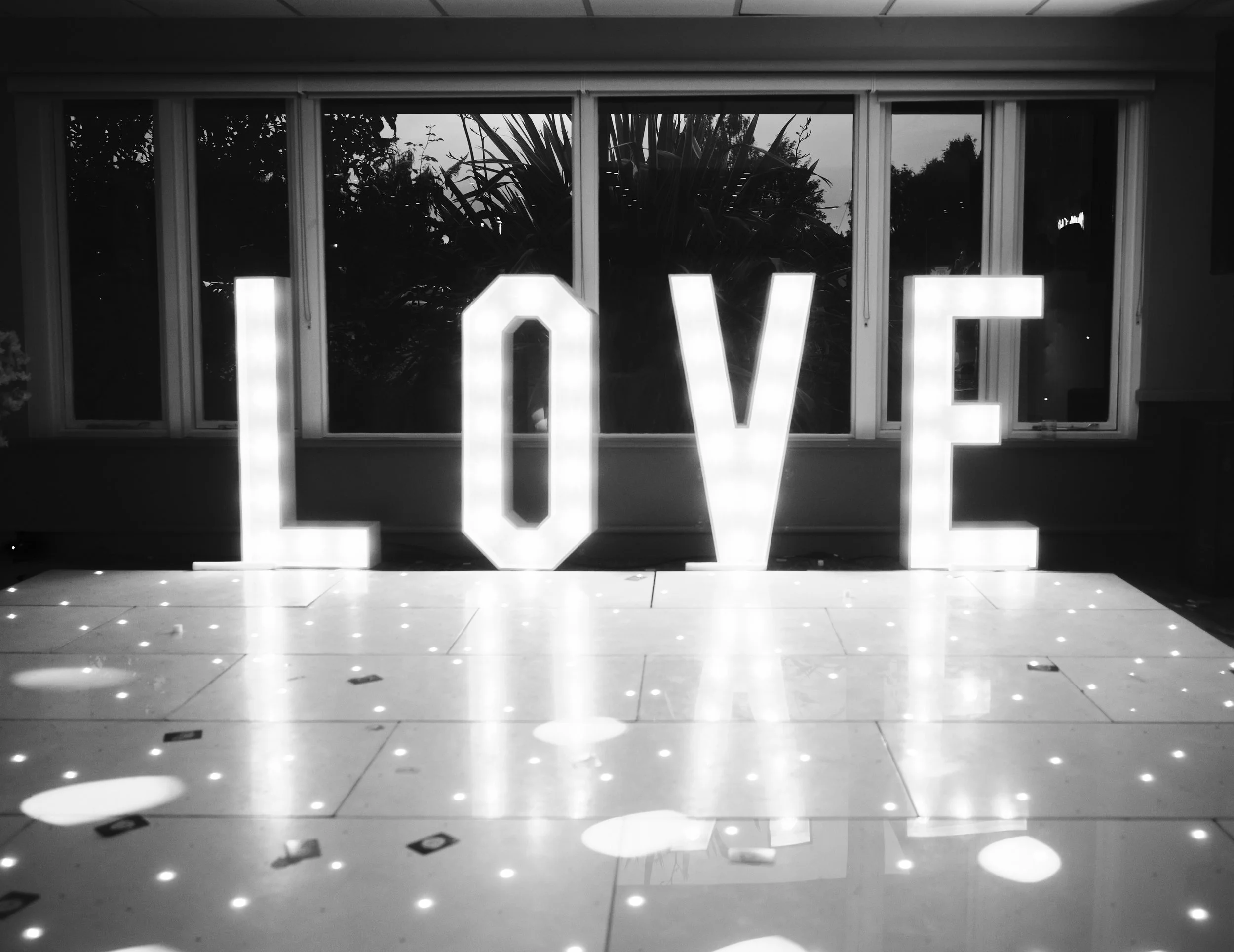 Illuminated decorative letters spelling 'LOVE' on a reflective floor, with a large window and trees outside in the background.
