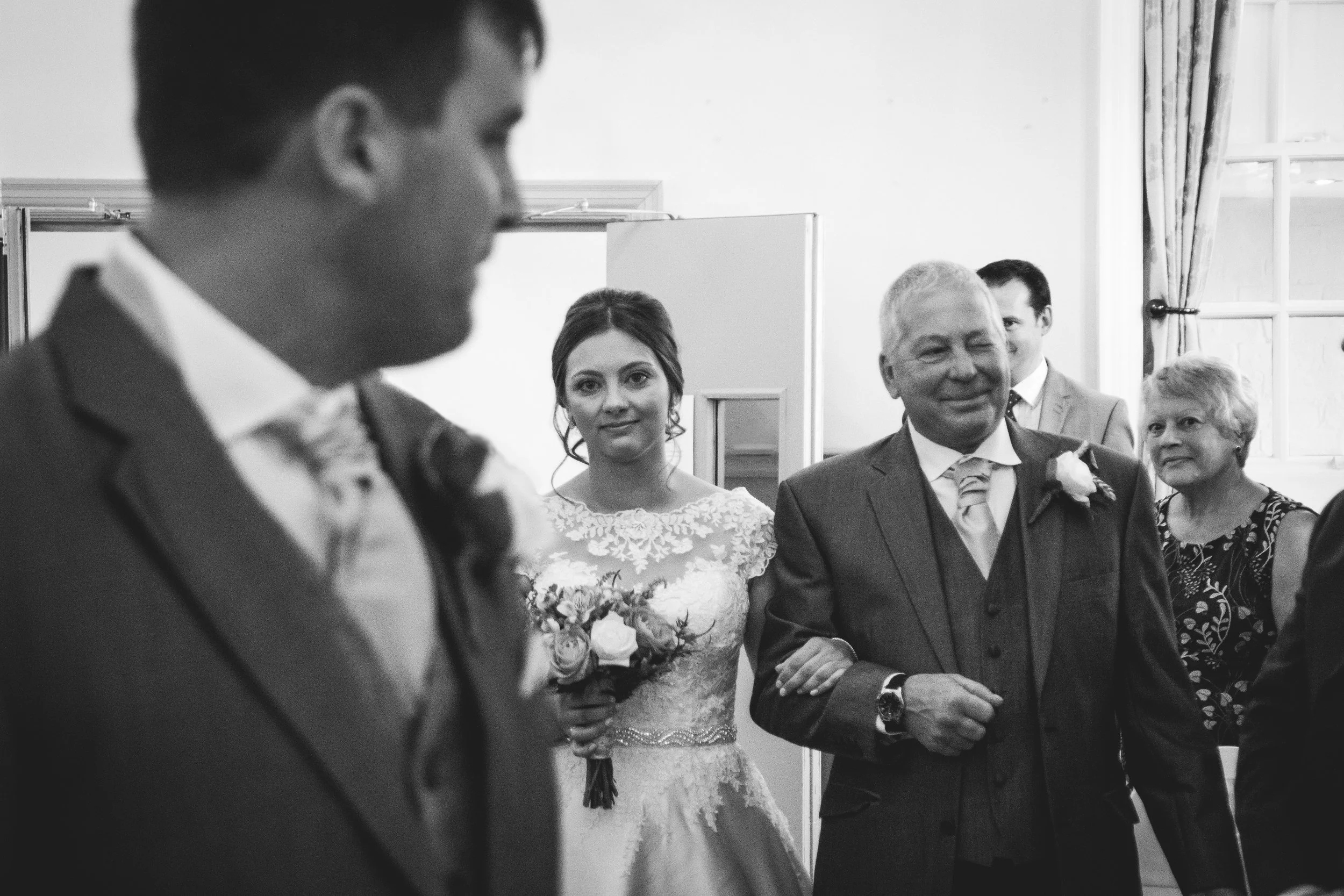 A bride in a lace wedding dress holding a bouquet, smiling, standing with an older man in a suit, surrounded by wedding guests in a room with a window and curtains.