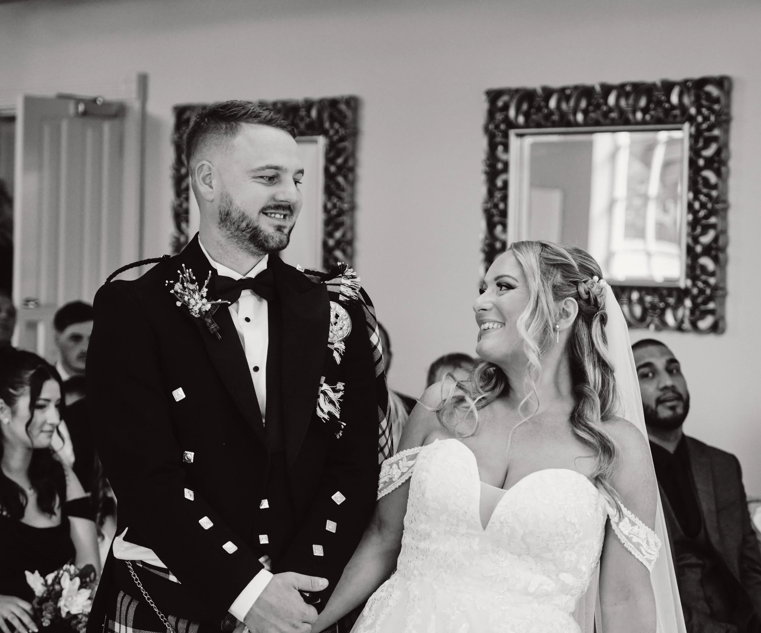 A bride and groom look at each other during their wedding ceremony. The groom wears a military uniform and the bride a lace wedding gown with a veil. Guests are seated in the background.