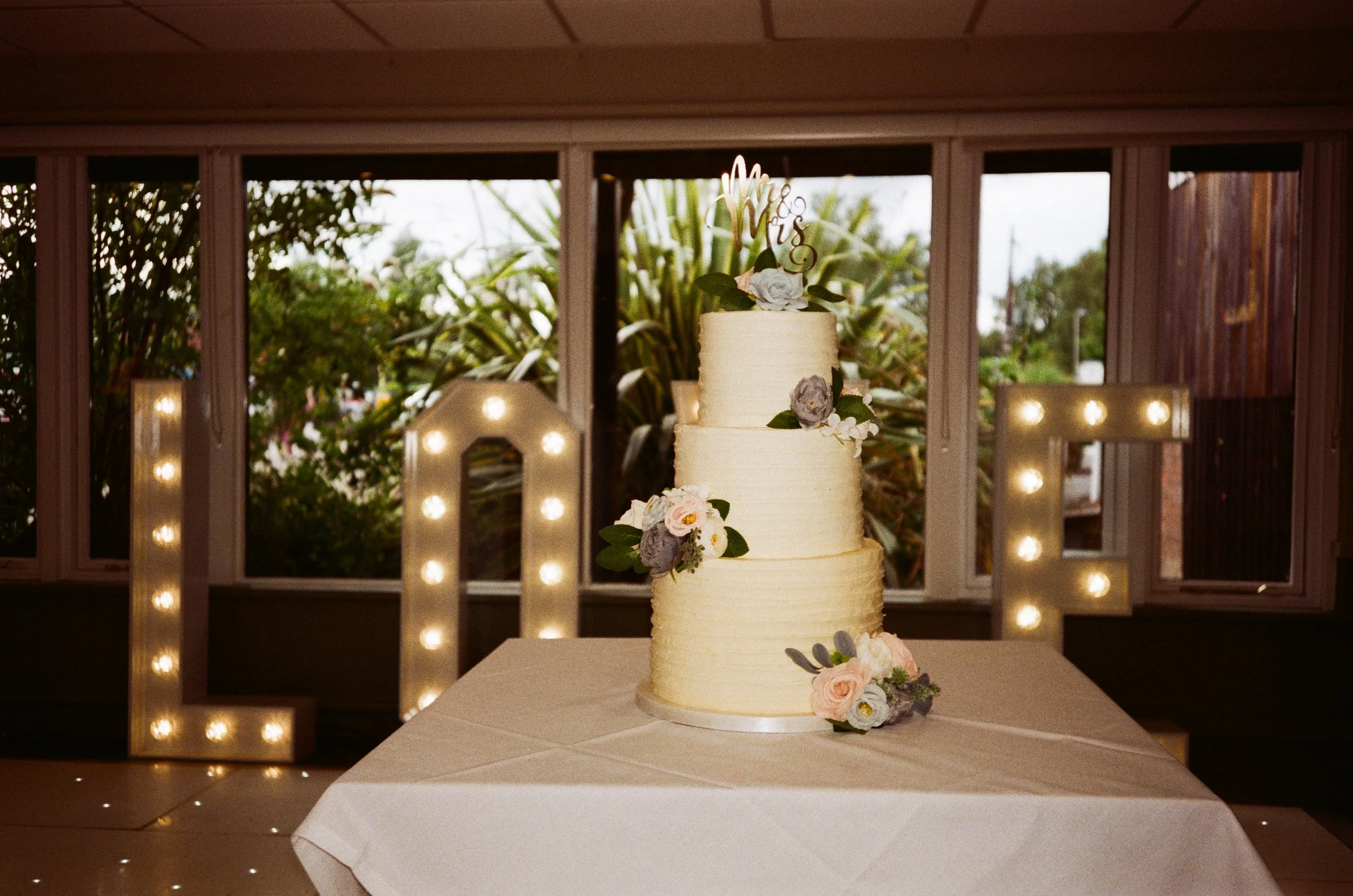 A multi-tiered wedding cake decorated with flowers, placed on a table with a white tablecloth, with large illuminated letters spelling 'LOVE' in the background.