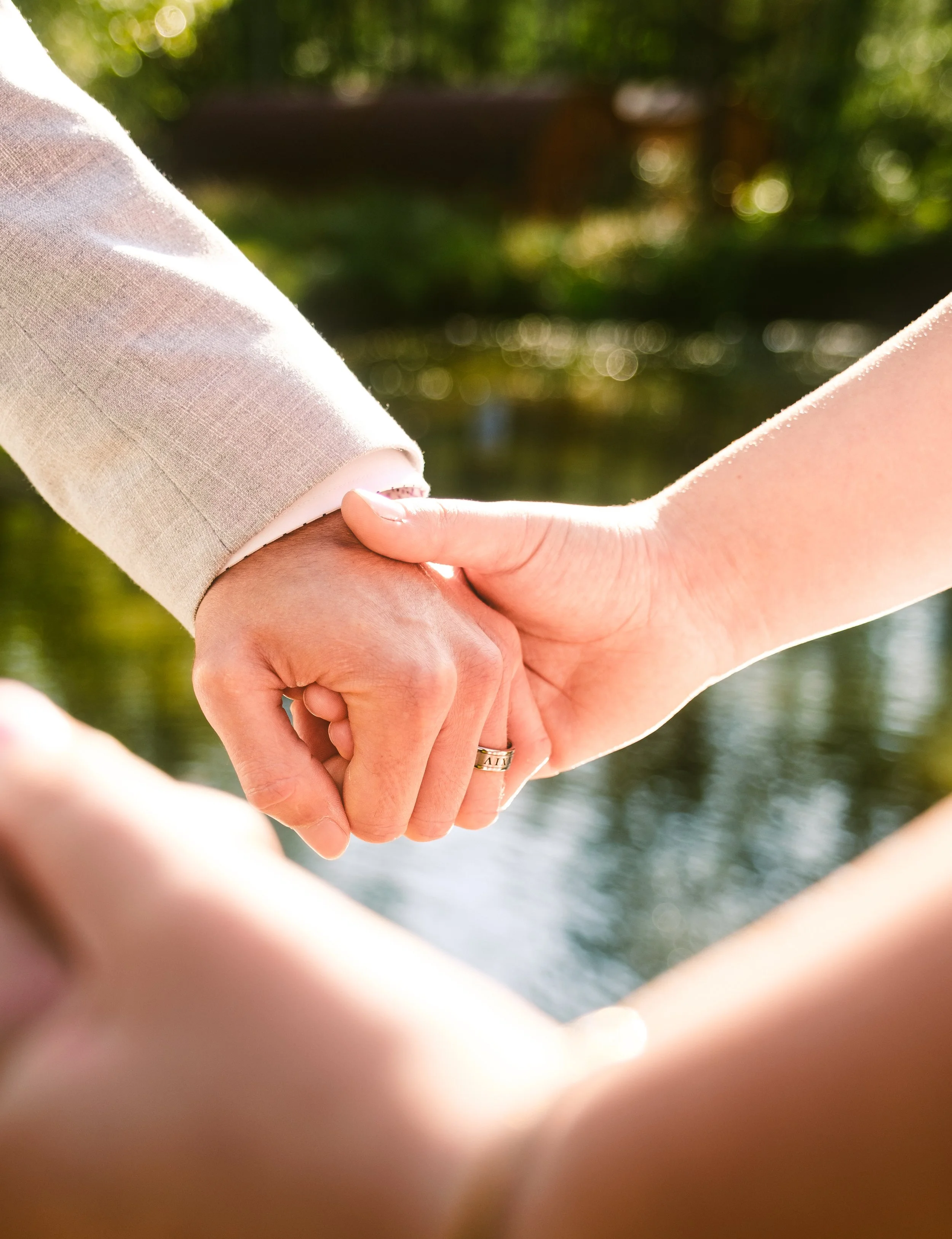 Close-up of two people holding hands near a body of water with blurred green trees in the background.