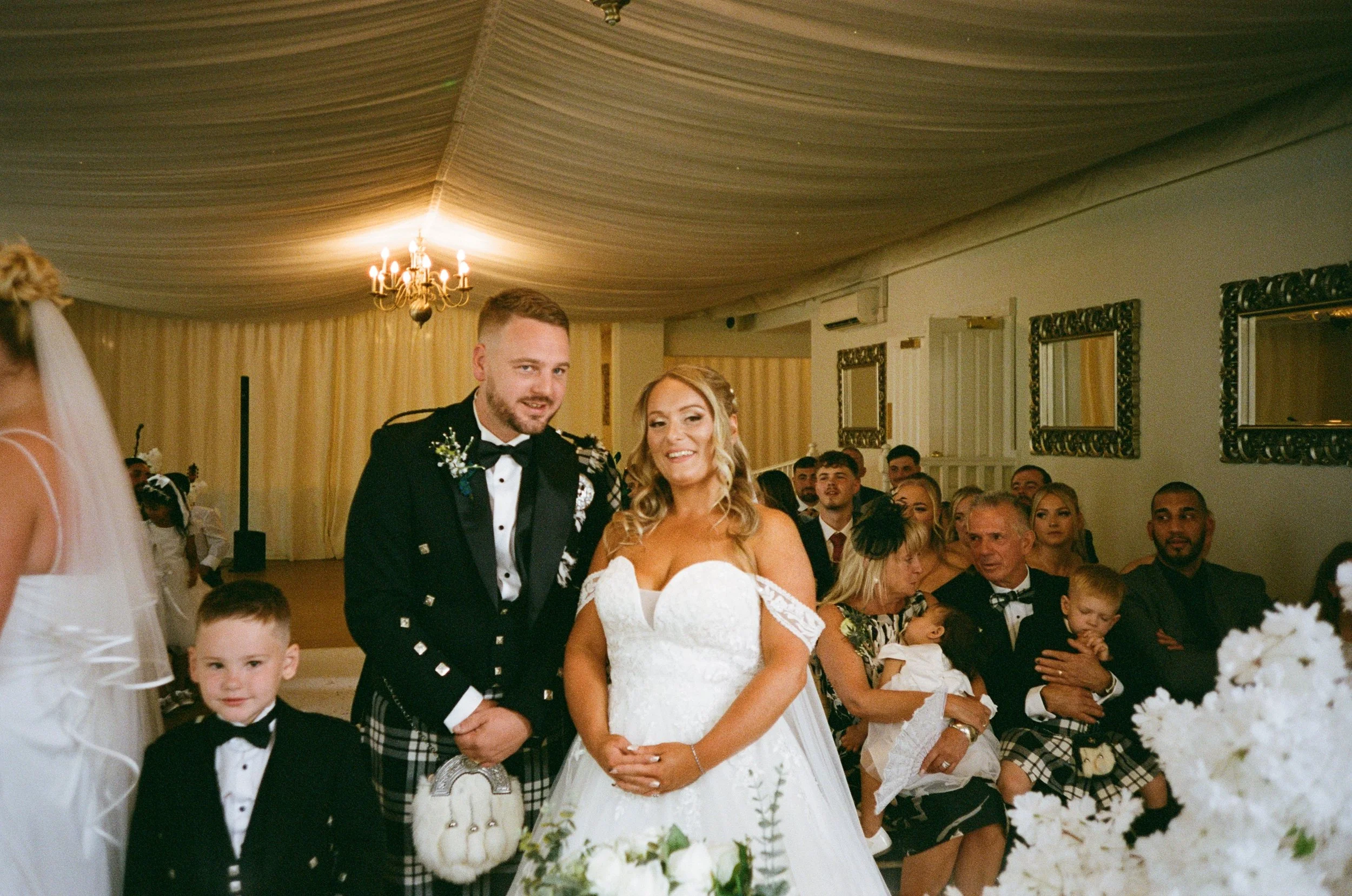 Bride and groom at their wedding ceremony, smiling and standing together. The bride wears a white dress with lace details, and the groom wears a traditional Scottish kilt with a black jacket and bow tie. Guests sit in the background, some dressed in