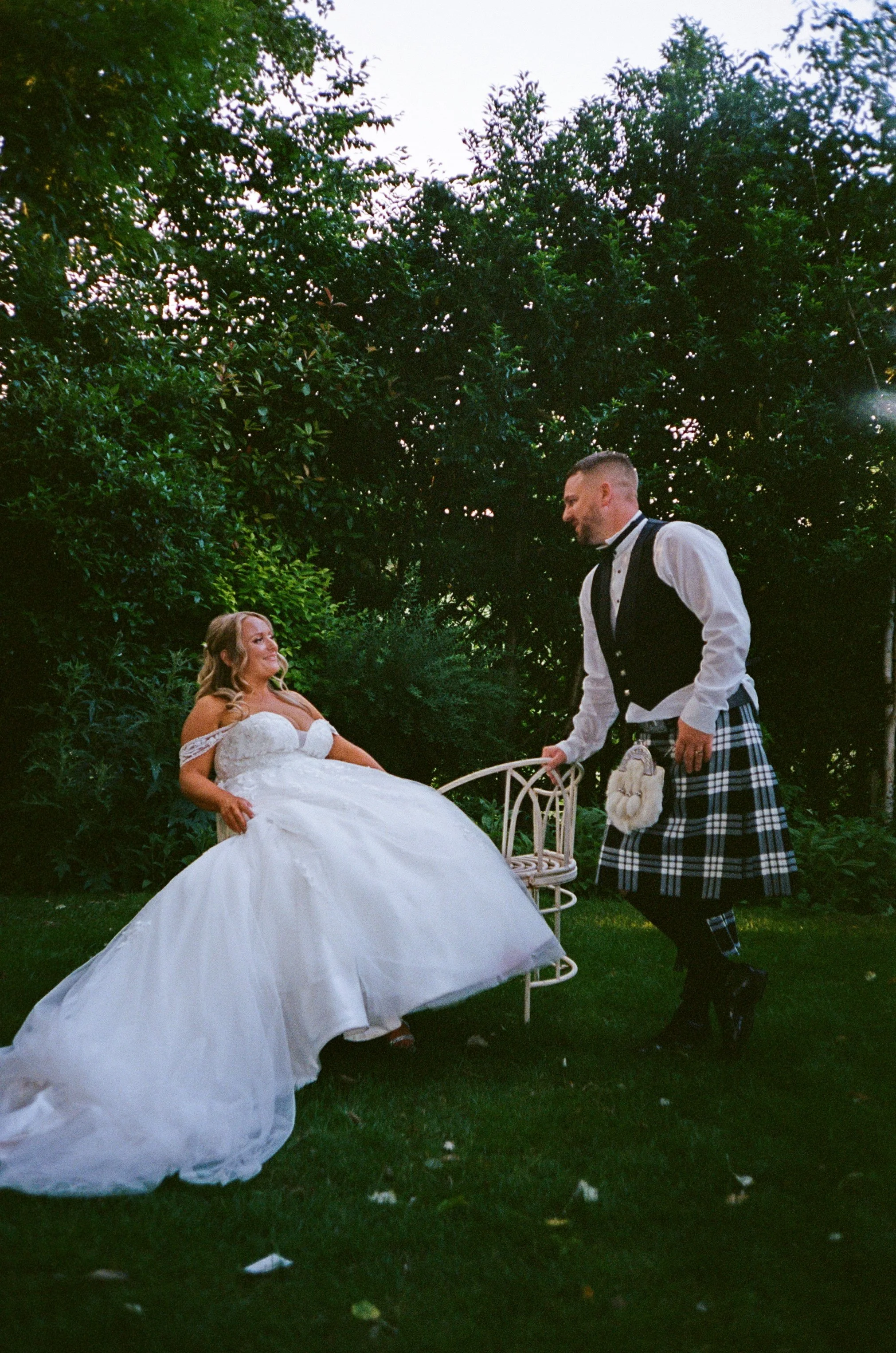 A bride in a white wedding gown sitting on a chair outdoors and a groom in Scottish attire standing and smiling at her amidst green foliage.