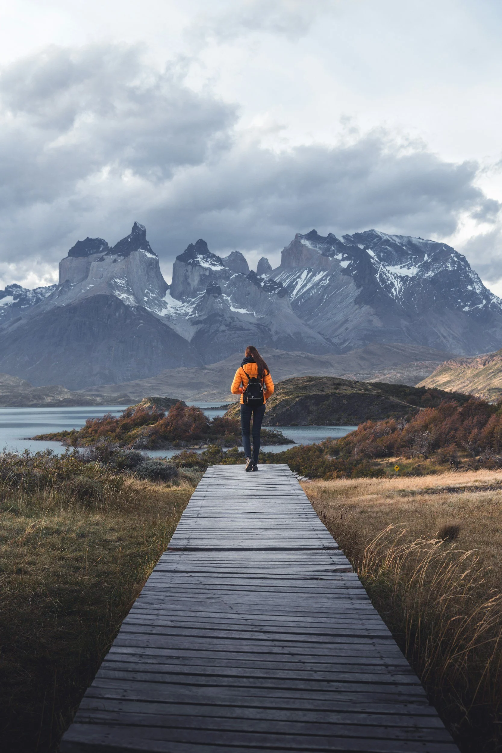 A person walking on a wooden path through a scenic landscape in Torres del Paine National Park in Chile with mountains, water, and cloudy sky in the background.