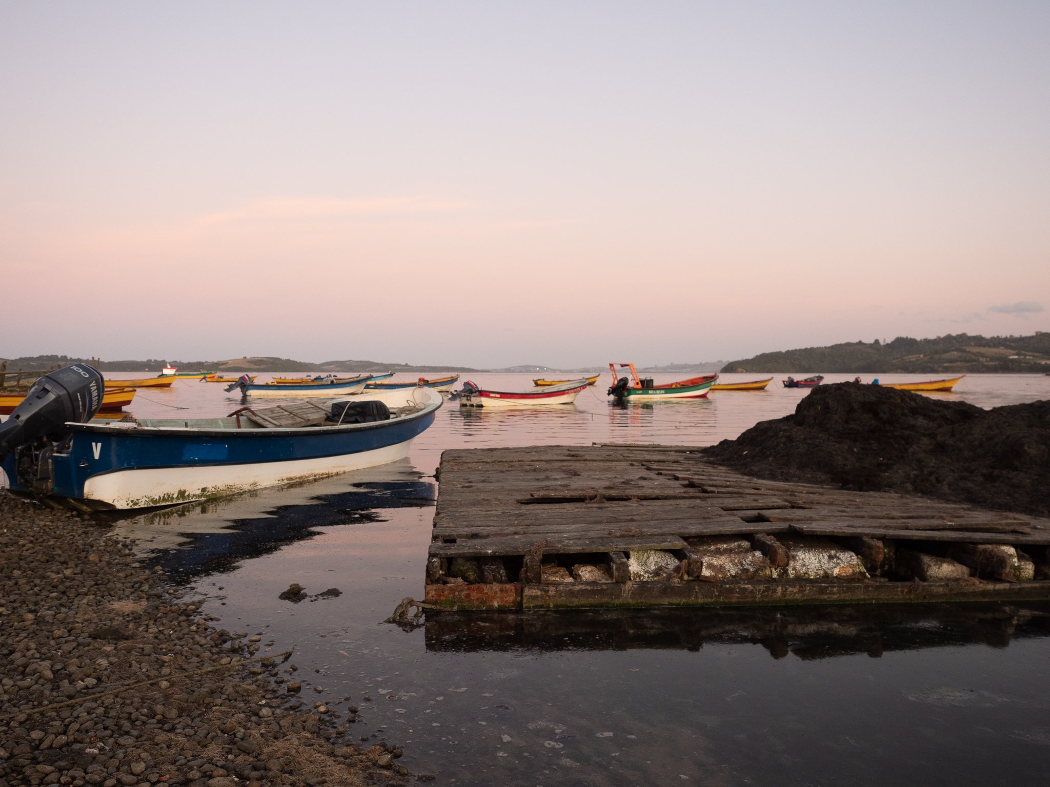 Small boats moored near a rocky shoreline with a wooden dock, calm water, and distant hills during sunset