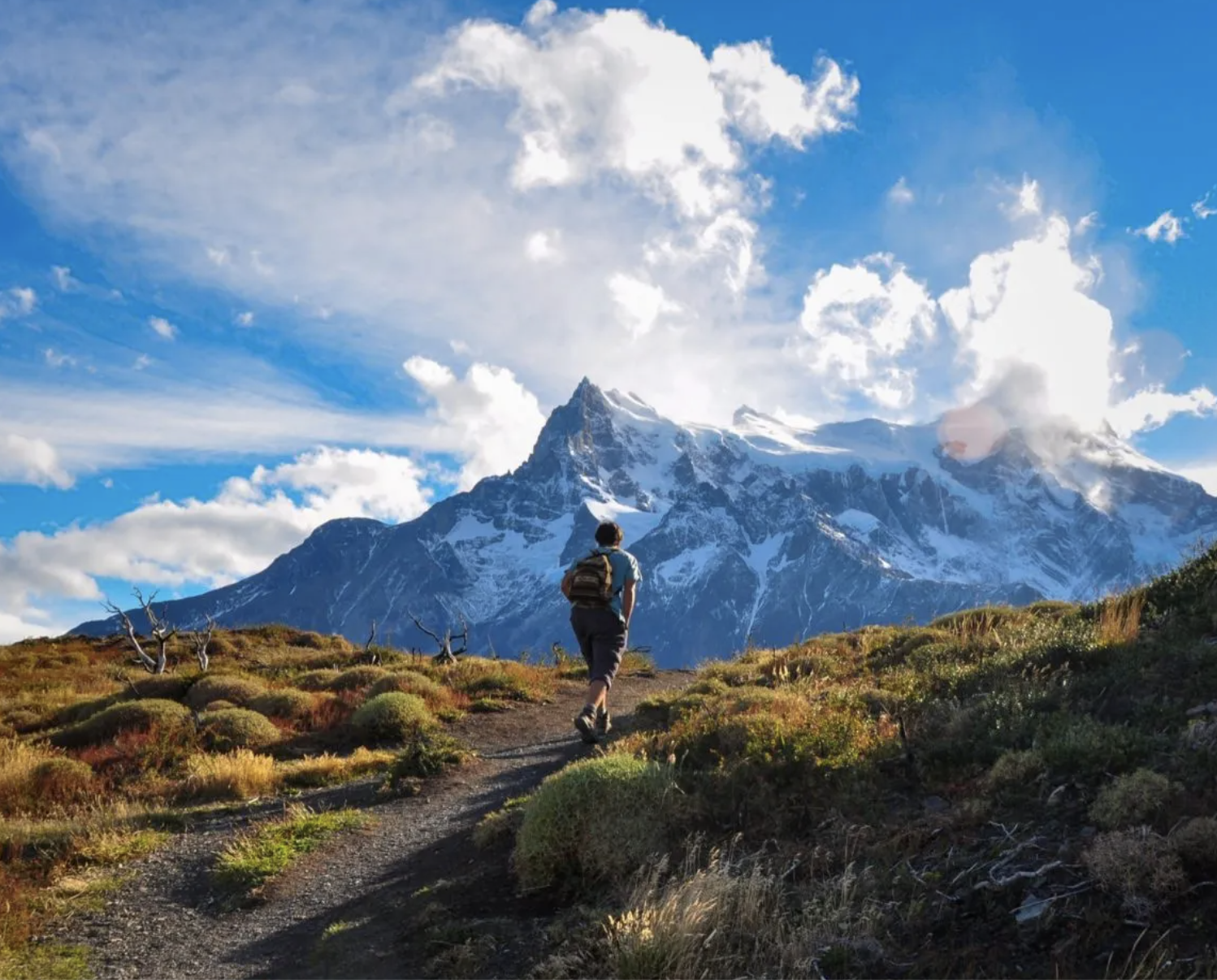 A person hiking on a trail through a grassy landscape with a snow-capped mountain in the background under a partly cloudy sky.