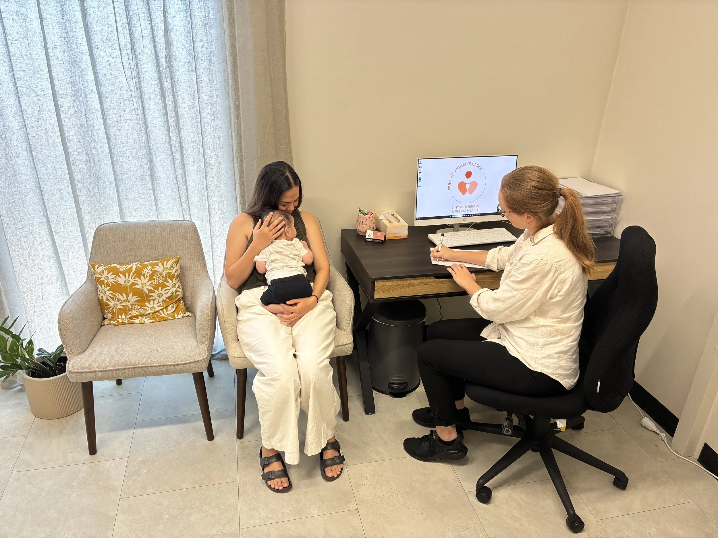 A woman sits on a beige armchair holding a baby, while a healthcare professional with glasses and a white coat writes notes at a desk. The scene takes place in a medical office with a monitor displaying a logo for the Territory Women's Clinic, focusing on physiotherapy and pelvic health, and a stack of papers on the desk.