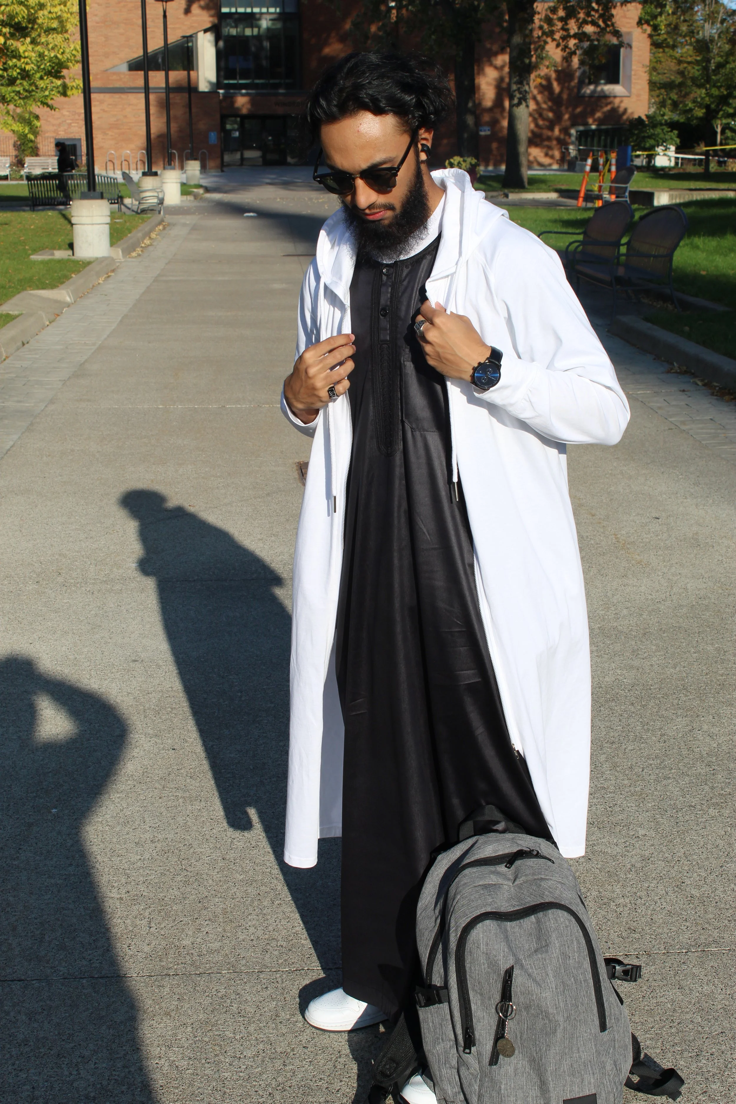 A man with a beard and sunglasses stands outdoors on a sidewalk, wearing a white hoodie thobe, holding a backpack in front of him, with a building and trees in the background.