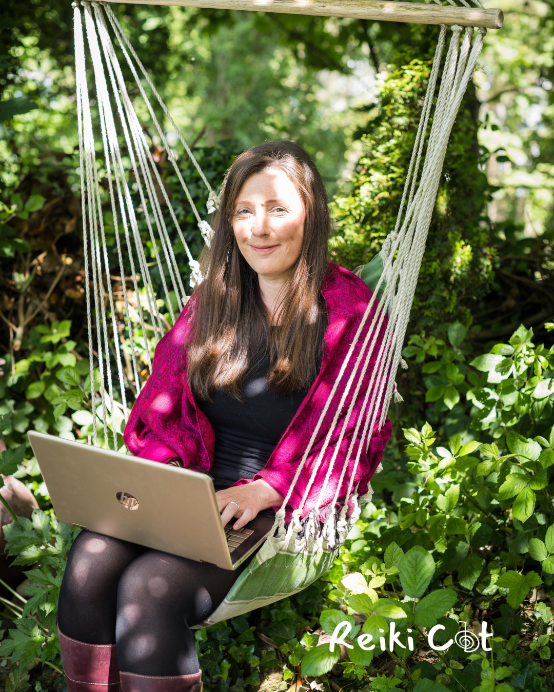 A woman with long brown hair sits in a hammock-style chair outside, working on a silver laptop. She is wearing a black top, a red shawl, black tights, and maroon boots. The background is filled with green foliage, and there is a logo that reads 'Reiki Cot' in the bottom right corner.