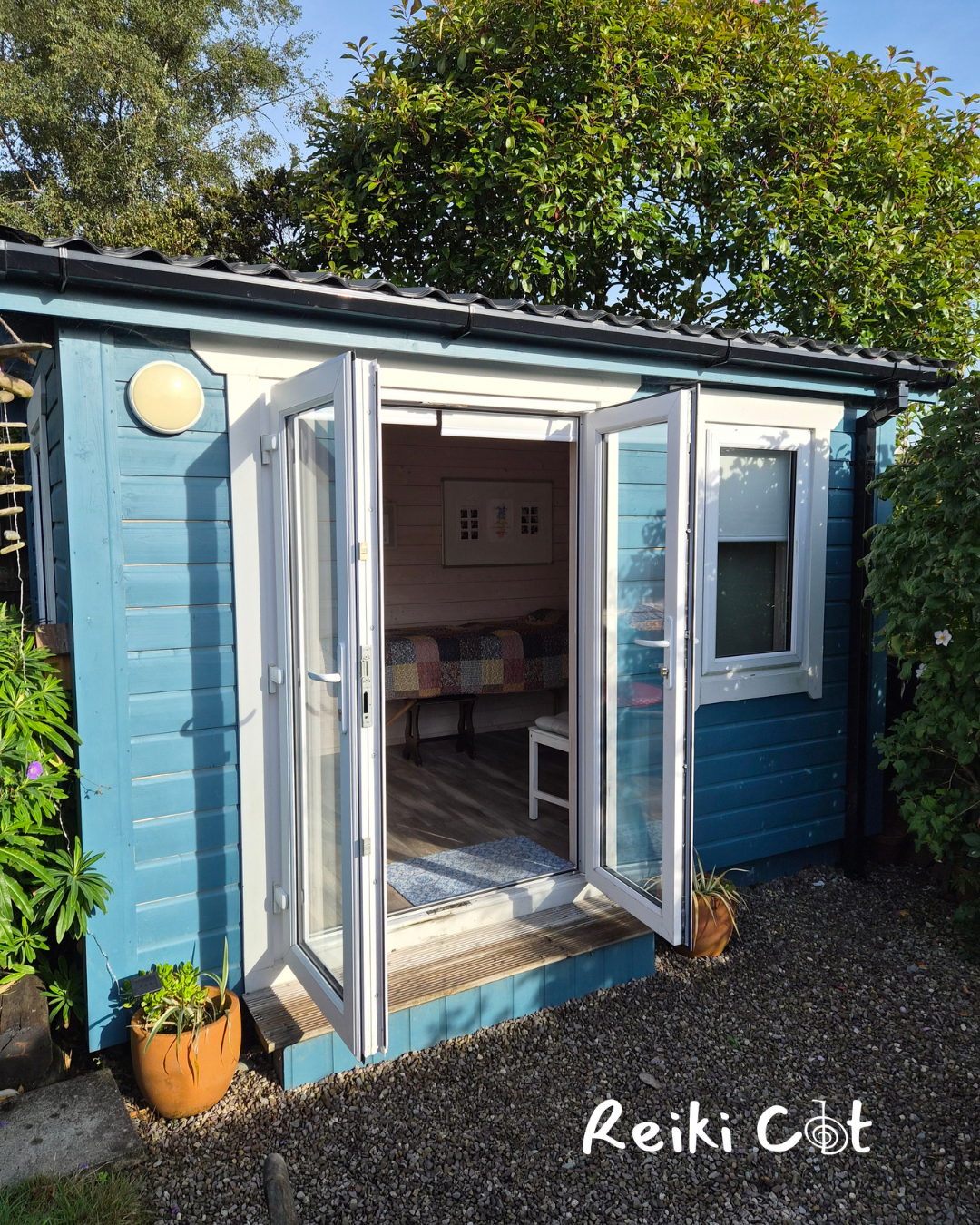 Small blue garden shed with open double glass doors, surrounded by greenery, with a potted plant outside on the gravel ground.