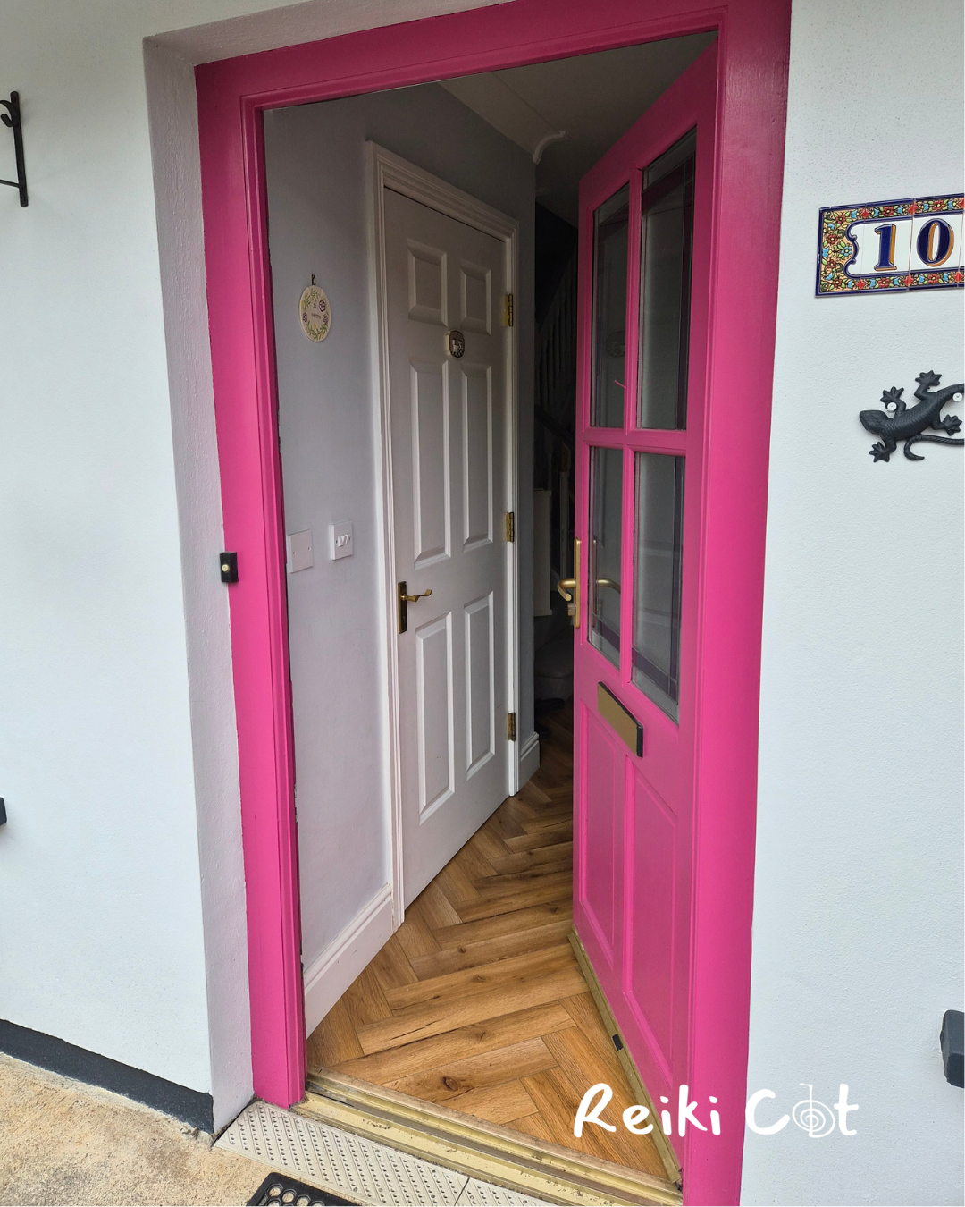 The image shows a front door entrance with a pink door frame and pink door with glass panels. Inside, there is a white interior door with a gold handle. The floor has wood flooring in a herringbone pattern. To the right of the entrance, there are decorative wall items, including a house number tile and a gecko-shaped ornament.