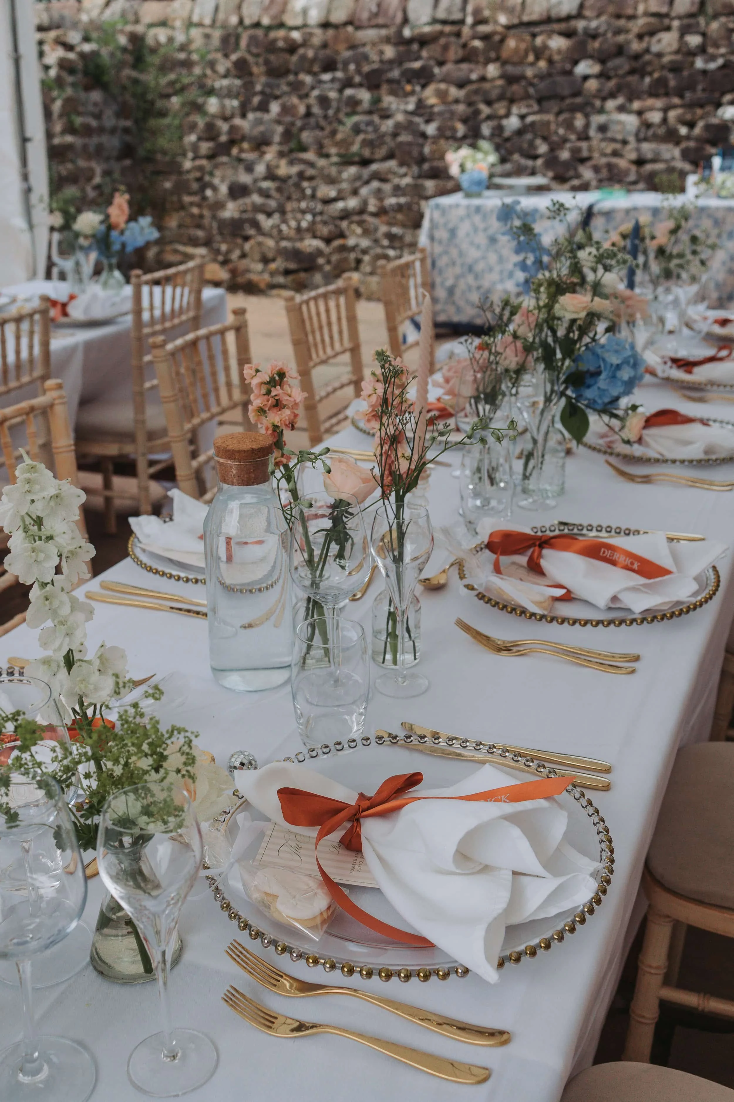 Elegant outdoor table setting with white tablecloths, floral centerpieces, gold flatware, napkins with orange ribbons, and glassware, against a stone wall background.