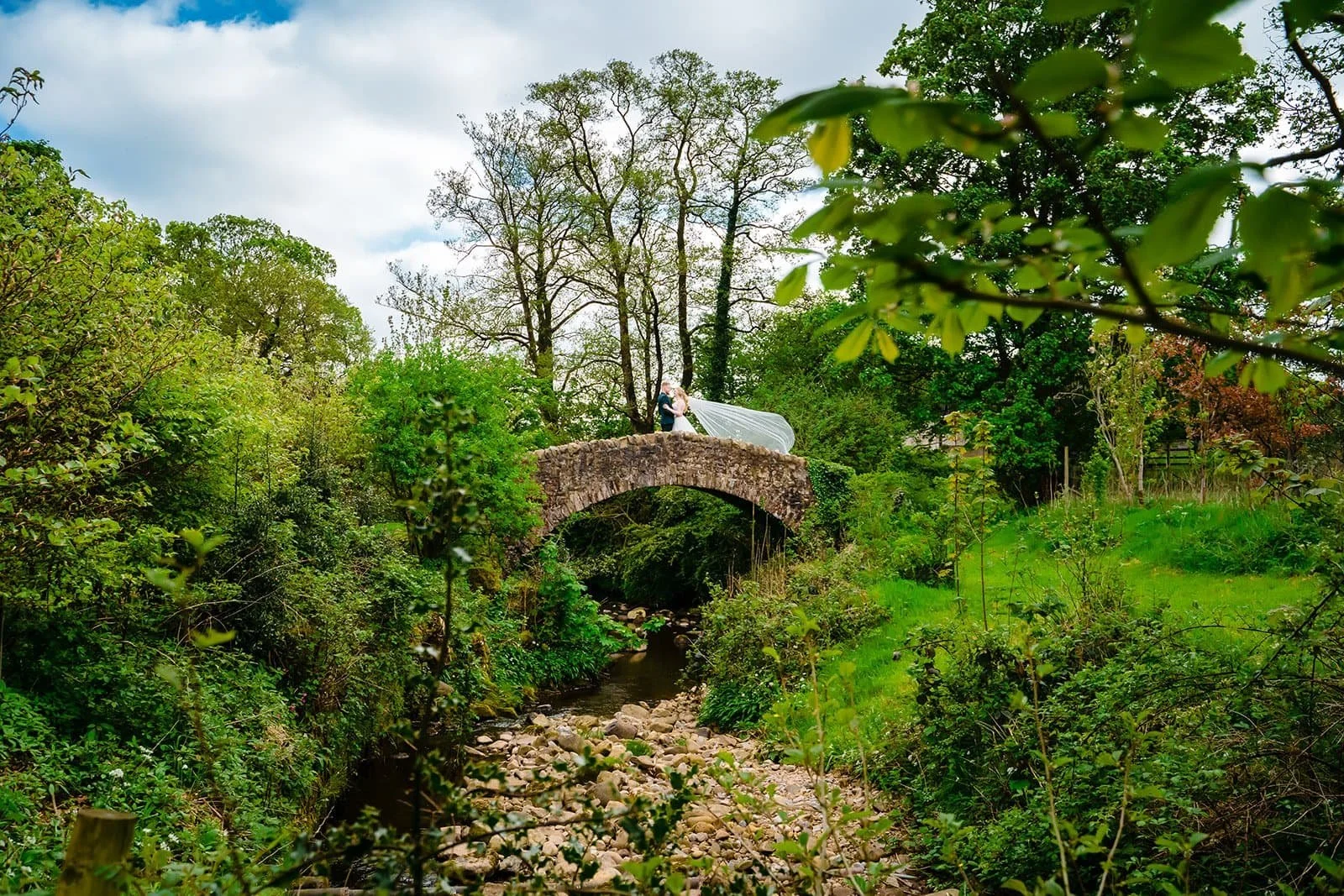 A bride and groom standing on a stone bridge over a babbling creek, surrounded by lush green trees and foliage, during a cloudy day.