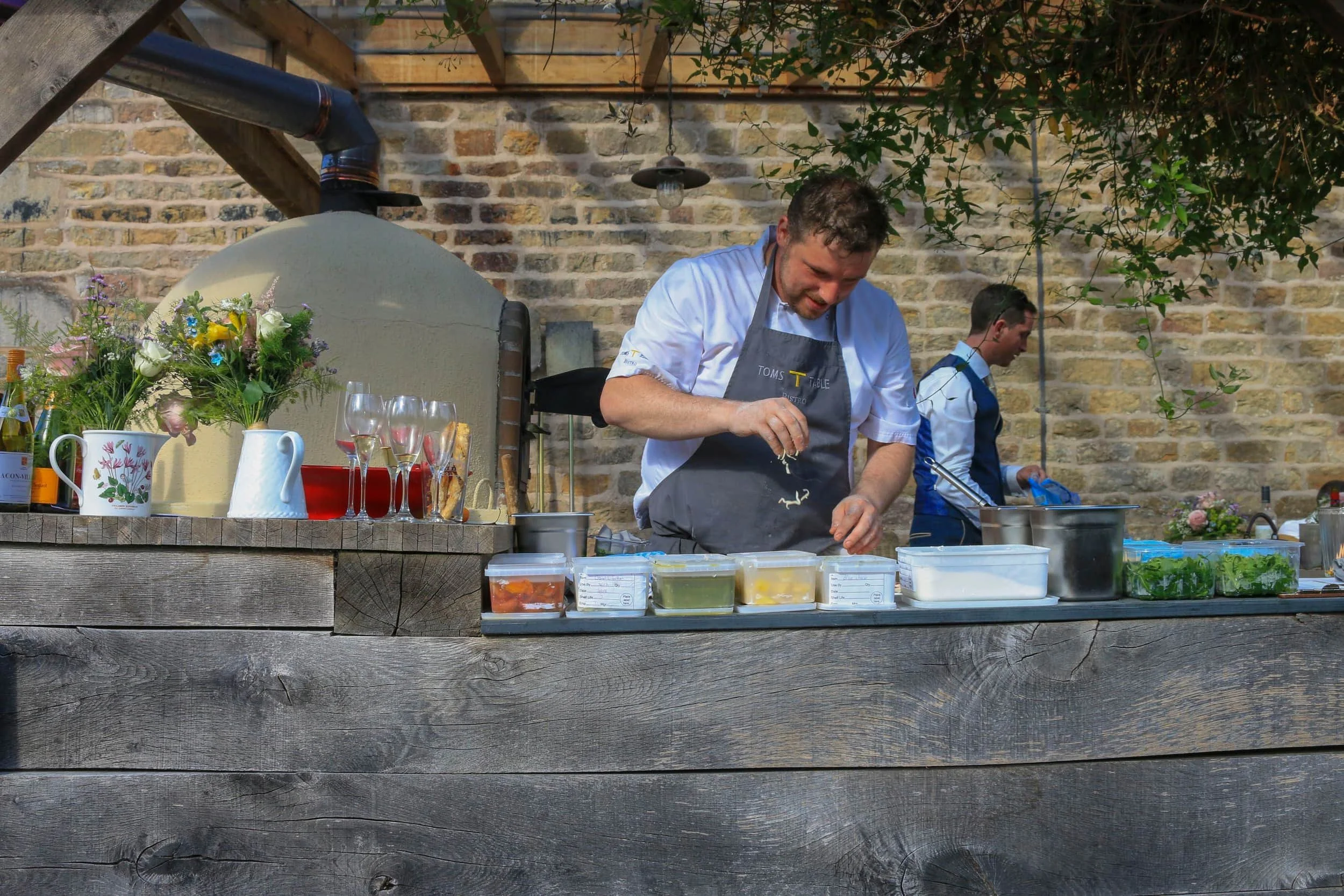 A chef wearing a gray apron and white shirt preparing food in an outdoor kitchen with a brick wall and greenery in the background, various kitchen utensils, ingredients, and flowers on the counter.