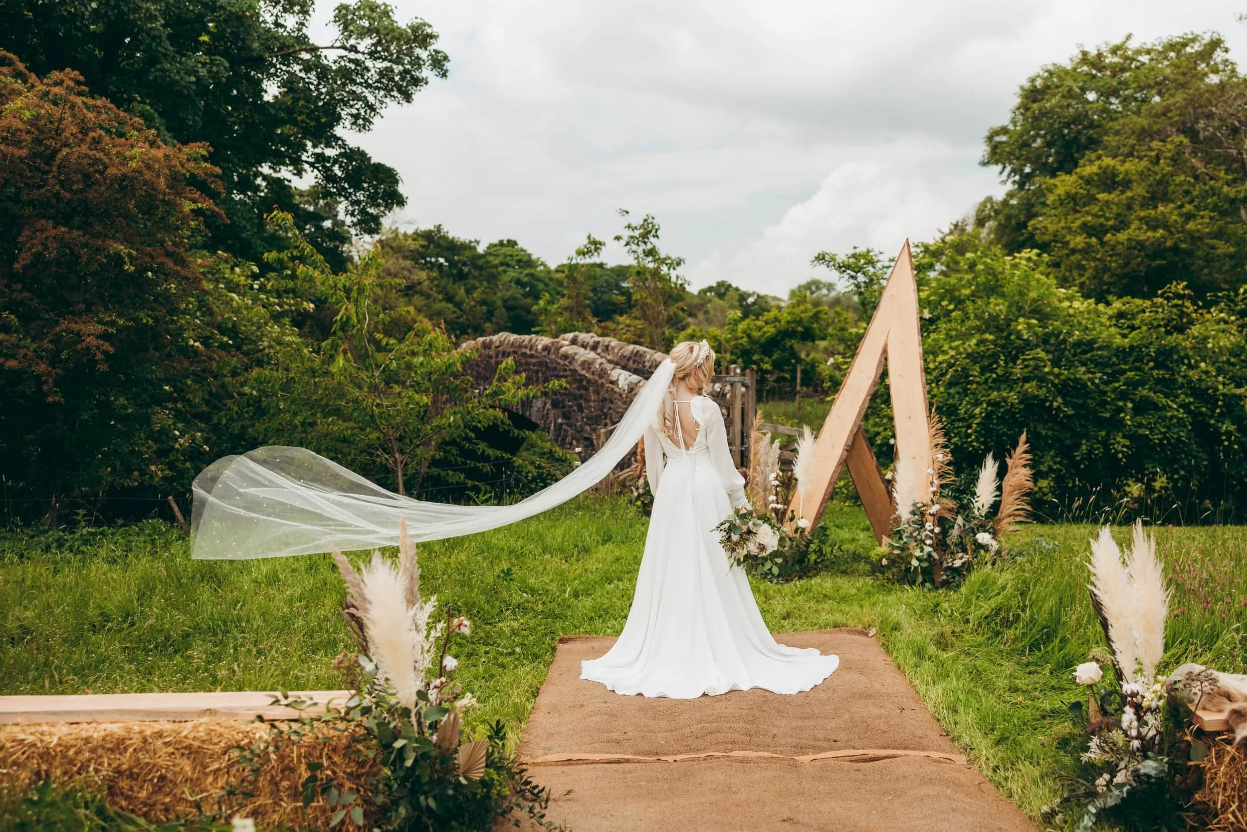 A bride in a white wedding dress with a long train and veil standing outdoors on a garden aisle with floral arrangements, surrounded by trees and greenery, near a wooden triangular arch.