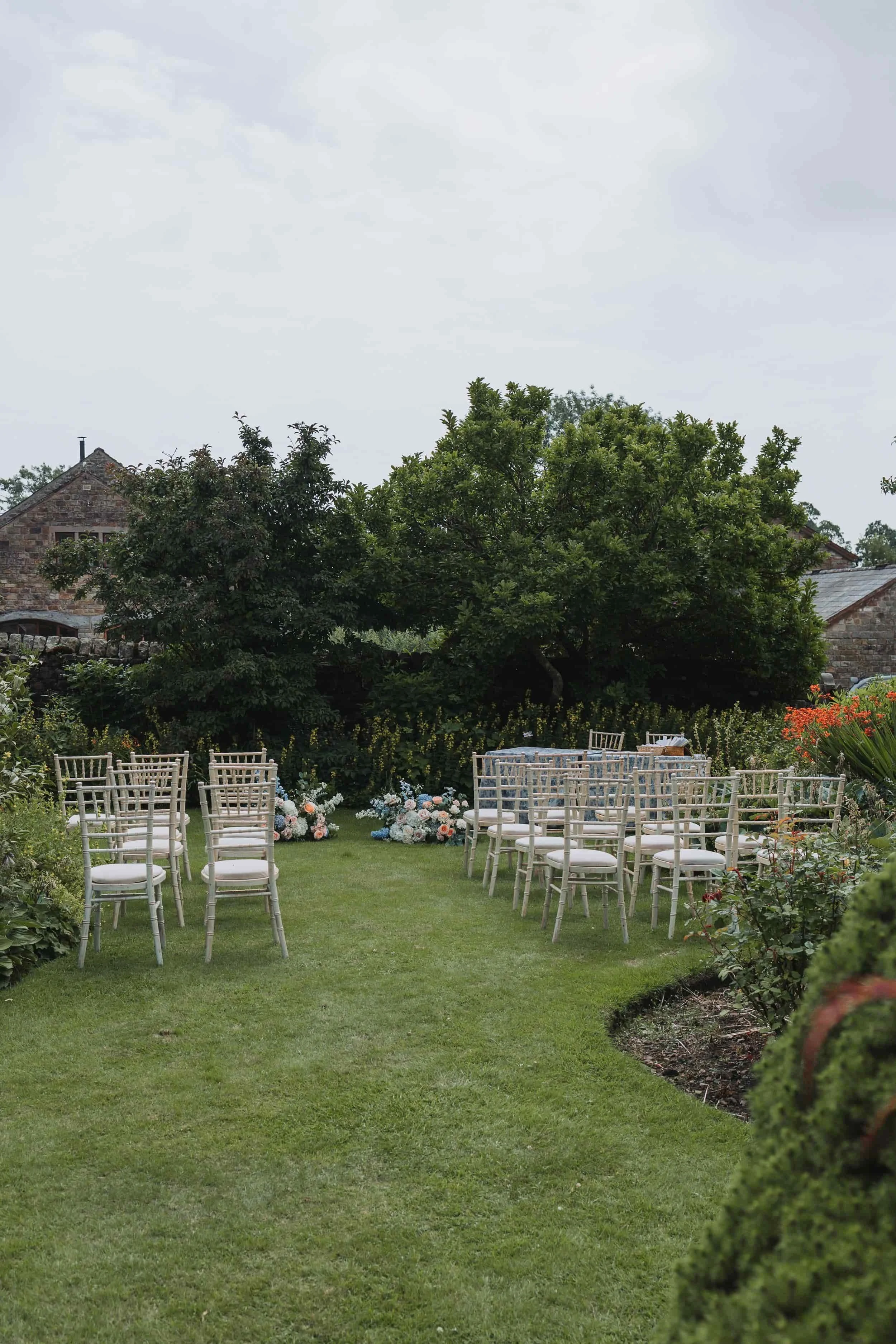Outdoor garden setting with white chairs arranged in rows on green grass, floral arrangements at the front, surrounded by bushes and trees, with stone houses and cloudy sky in the background.