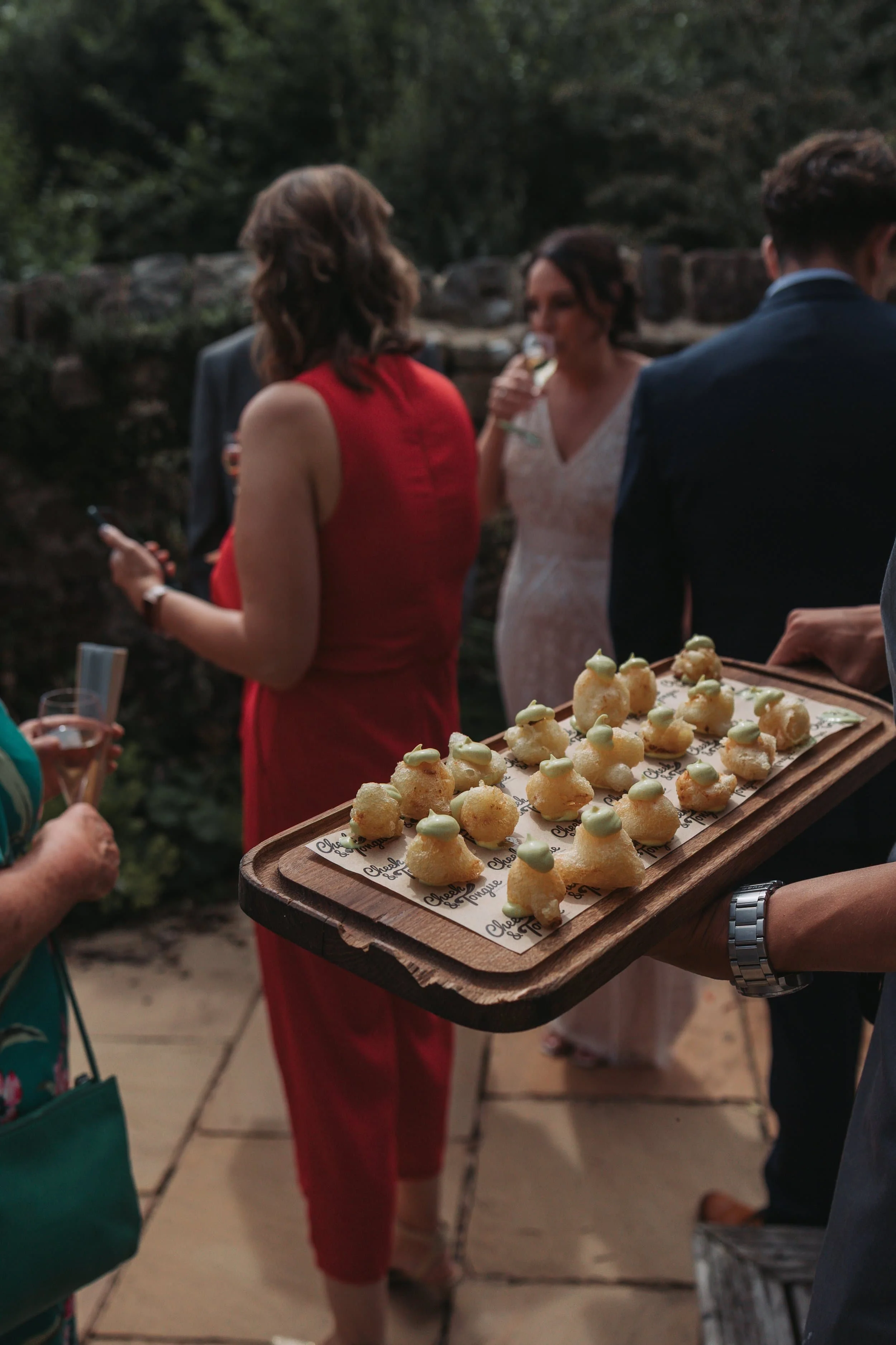 A person holding a tray with small pastries or appetizers during an outdoor gathering, with other guests in the background.