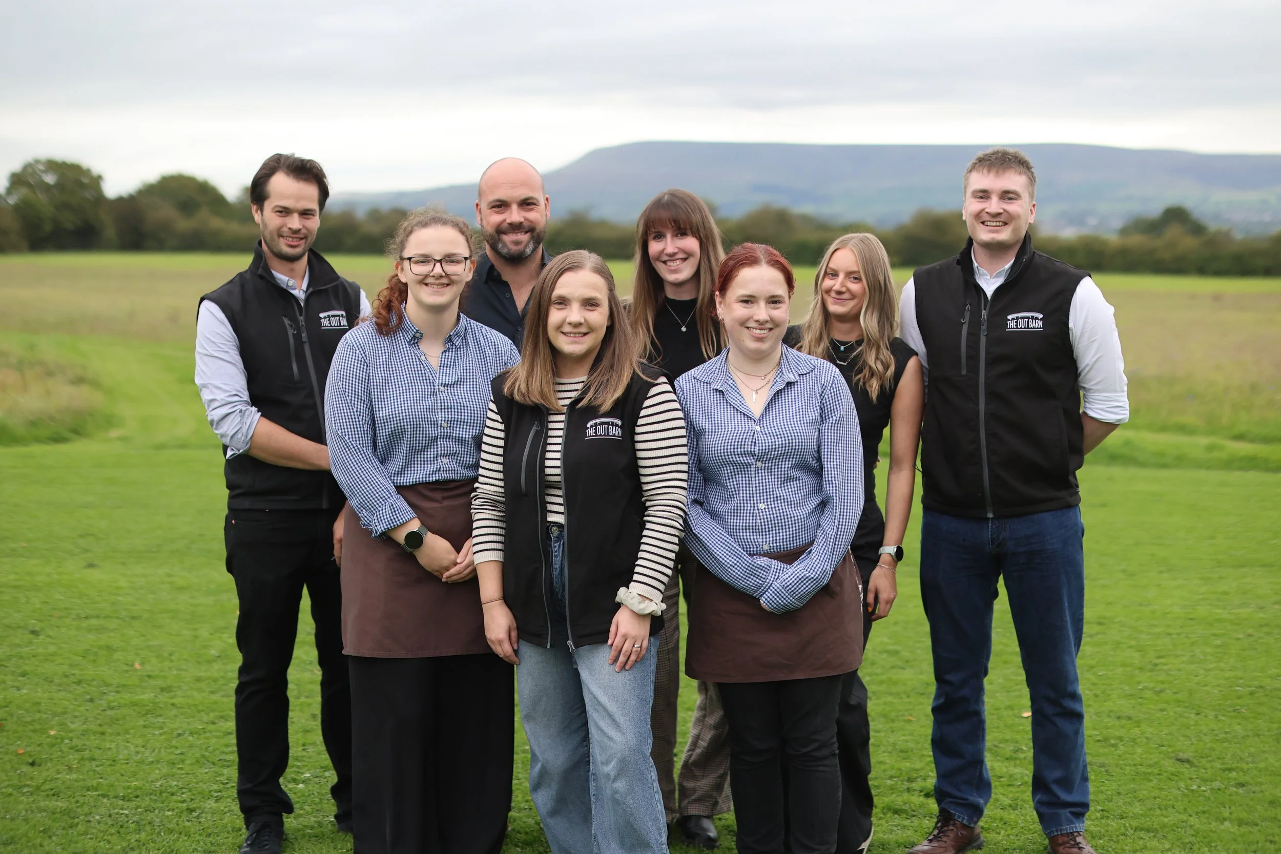 A group of nine smiling people standing outdoors on grass with a scenic background of trees and distant hills under cloudy skies.