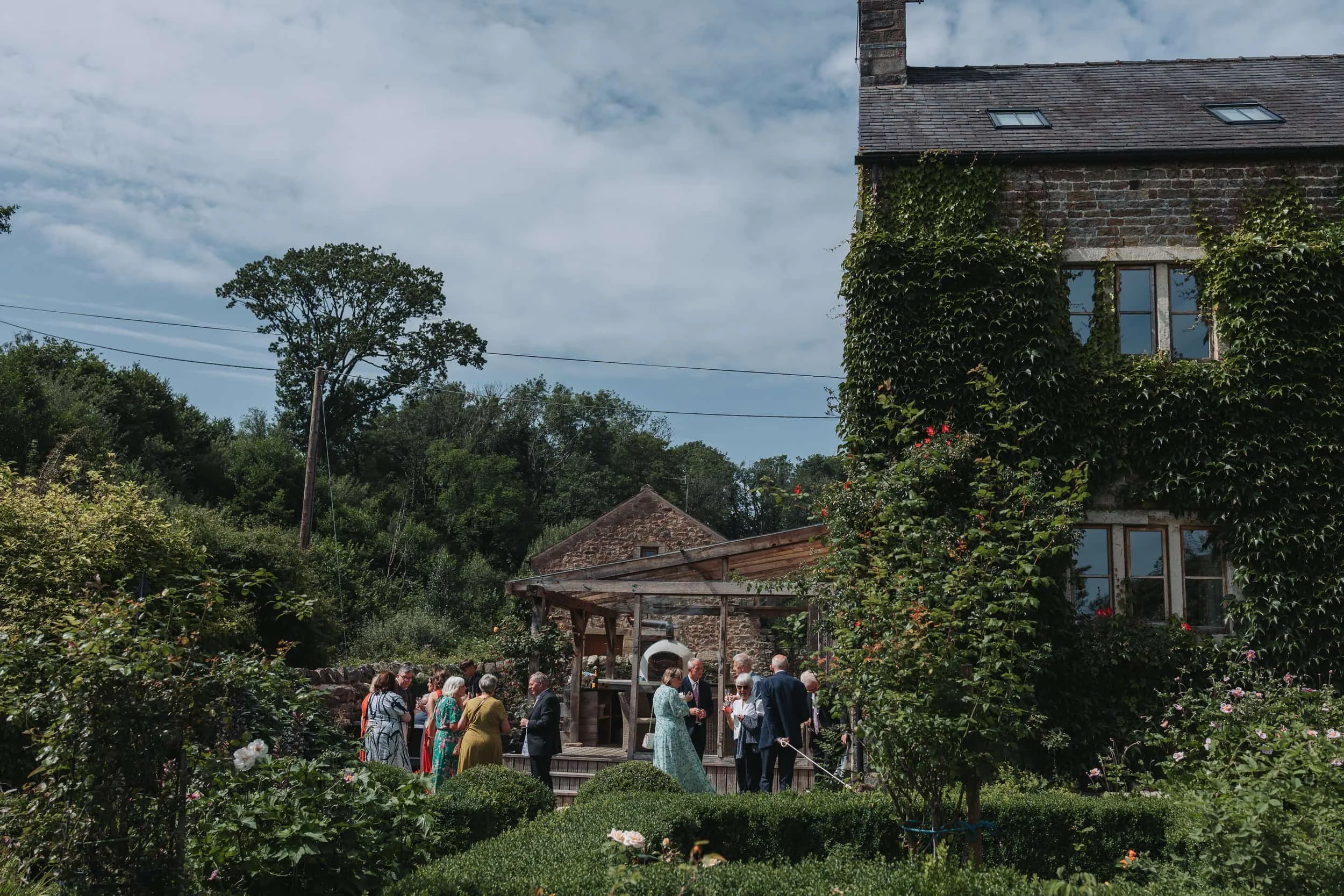A group of people gathered outdoors in a garden with lush greenery and flowers, near a stone and brick house covered in vines, with trees and a cloudy sky in the background.